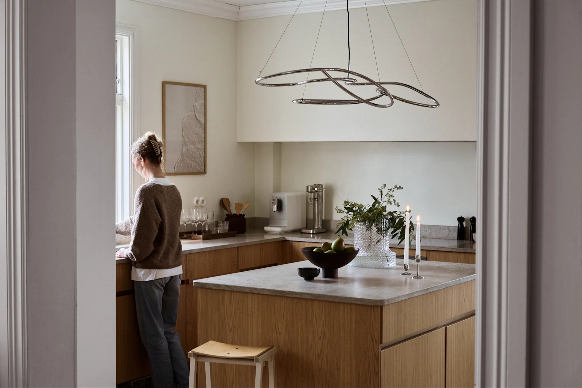 Woman looking out a window in a bright modern kitchen with light wood cabinets, gray countertops, and a sculptural pendant light.
