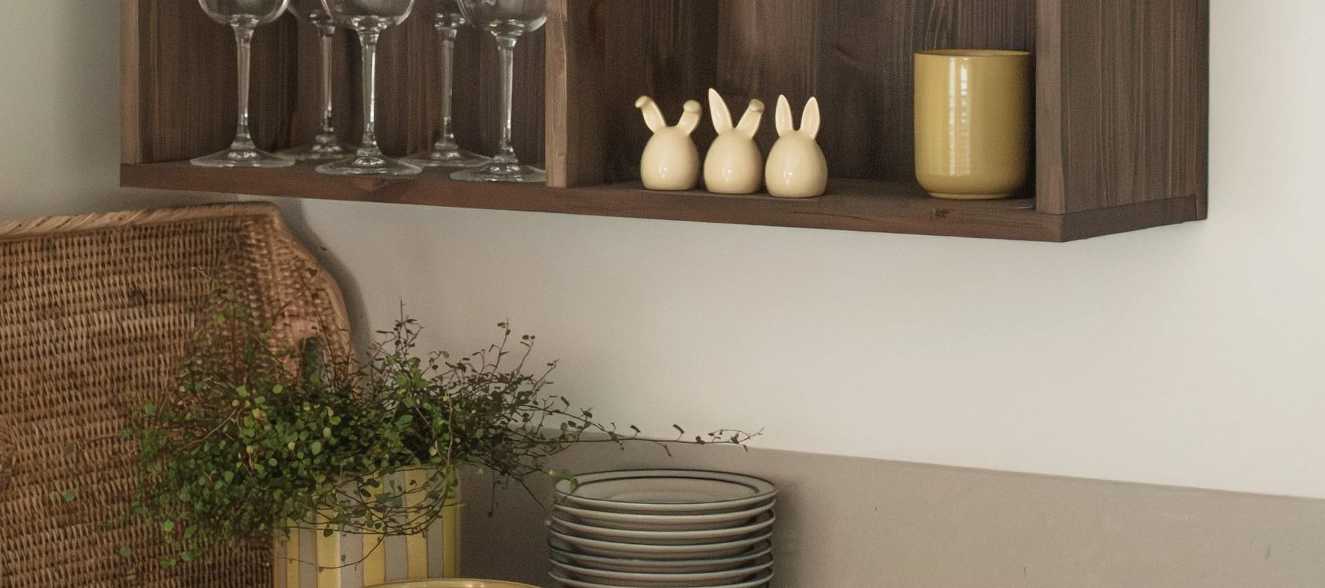 A wooden shelf holds glasses, three rabbit figurines, and a yellow mug, with a wicker basket and plants below.