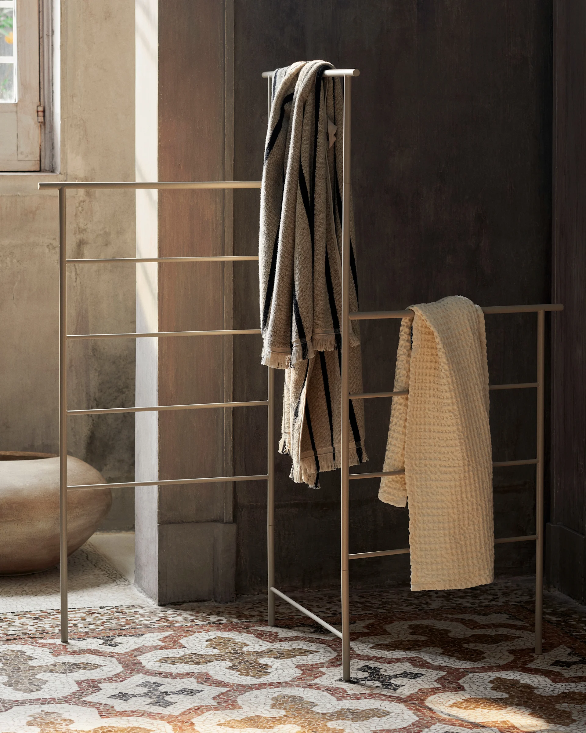 A freestanding metal towel rack holds a striped towel and an off-white textured towel in a room with a mosaic floor.