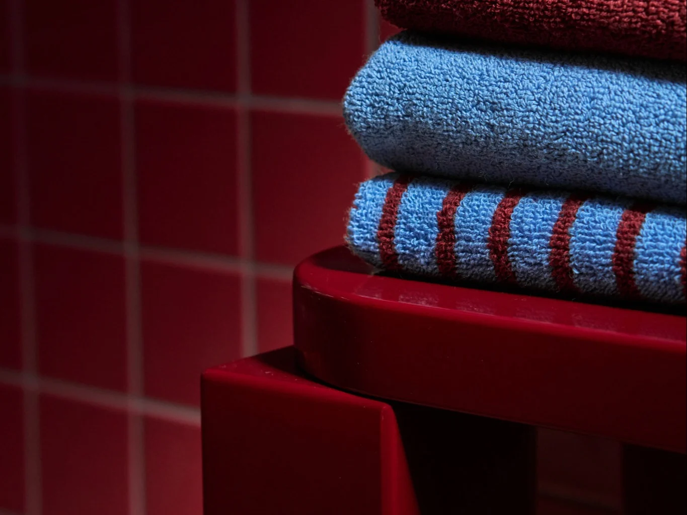 Stack of three folded towels: dark red, light blue, and blue with red stripes, on a red surface.