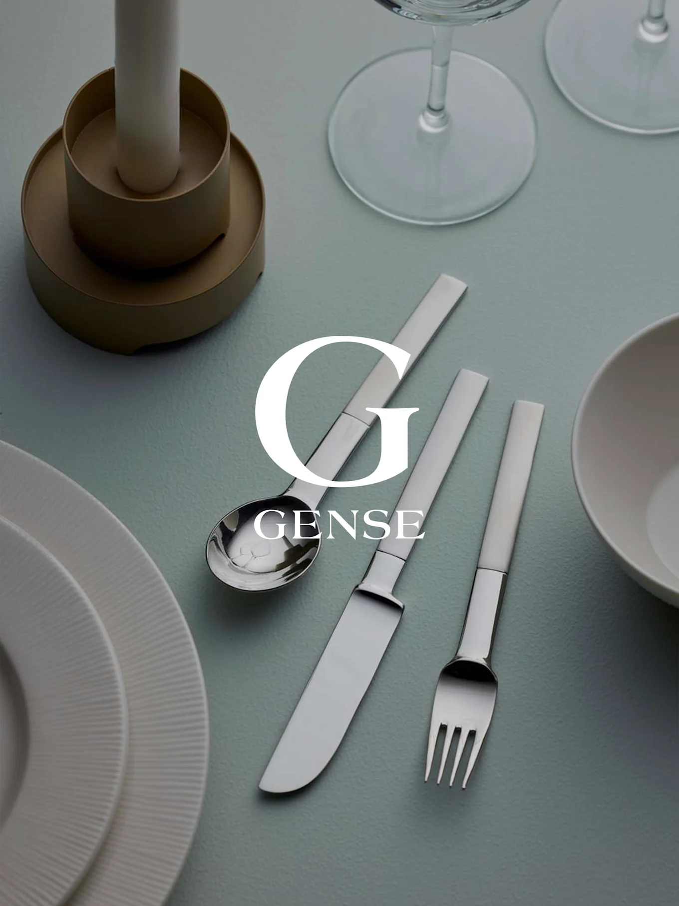 A table setting with Gense silver cutlery set (spoon, knife, fork), white plates, a candle, and glasses on a light blue surface.