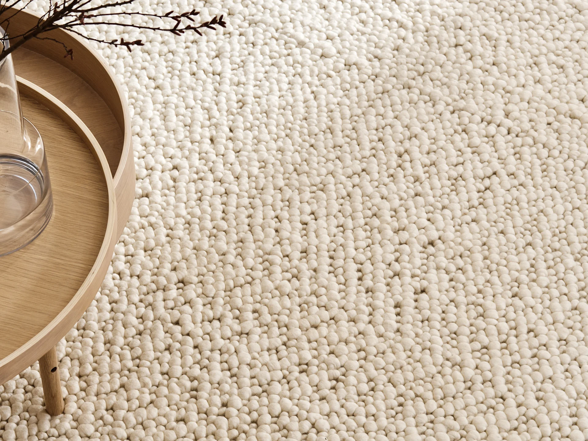 Close-up of a cream bobble rug with a light wooden side table and branches in a vase.