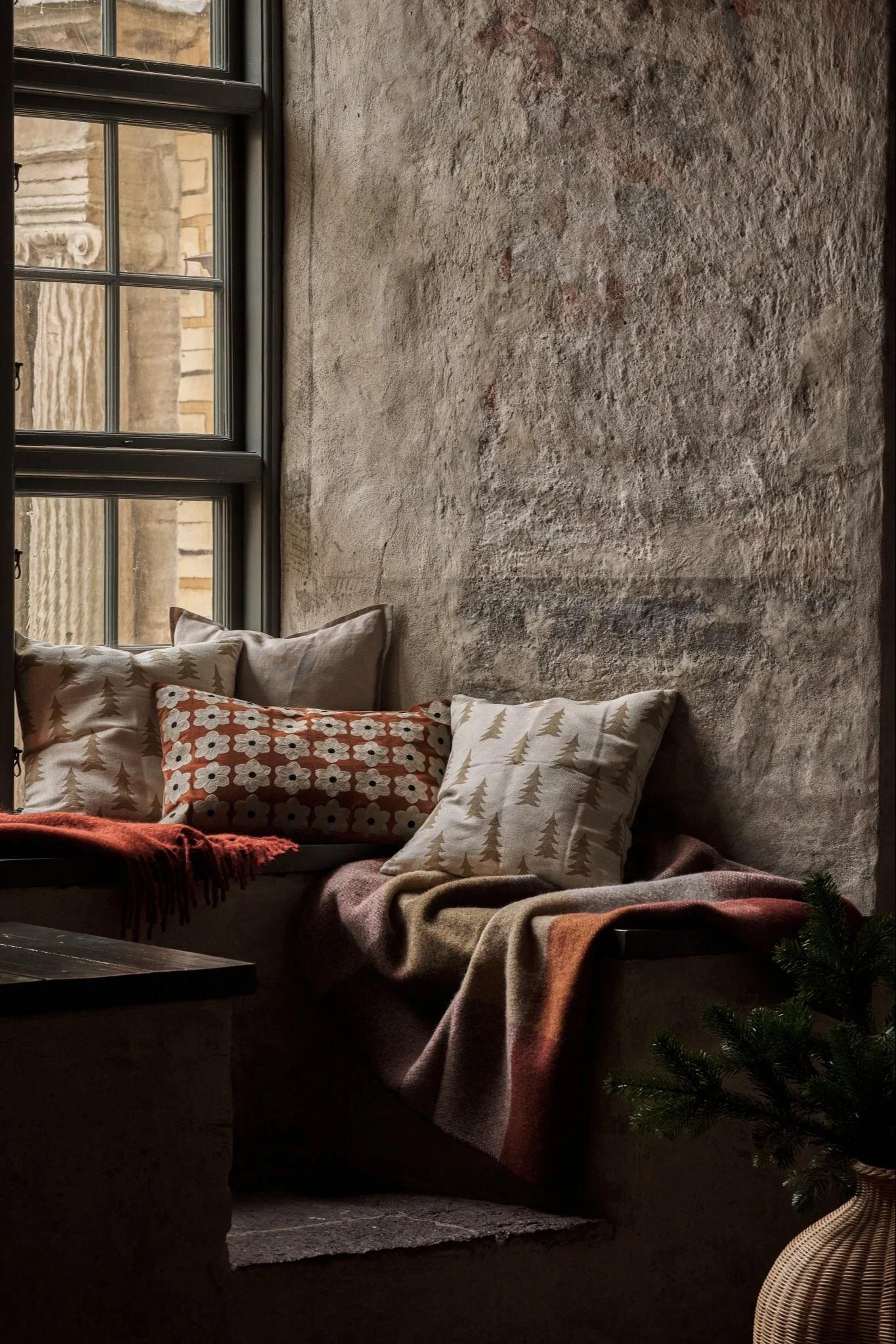 Cozy window seat with patterned pillows, warm blankets, and a pine branch next to a rustic wall.
