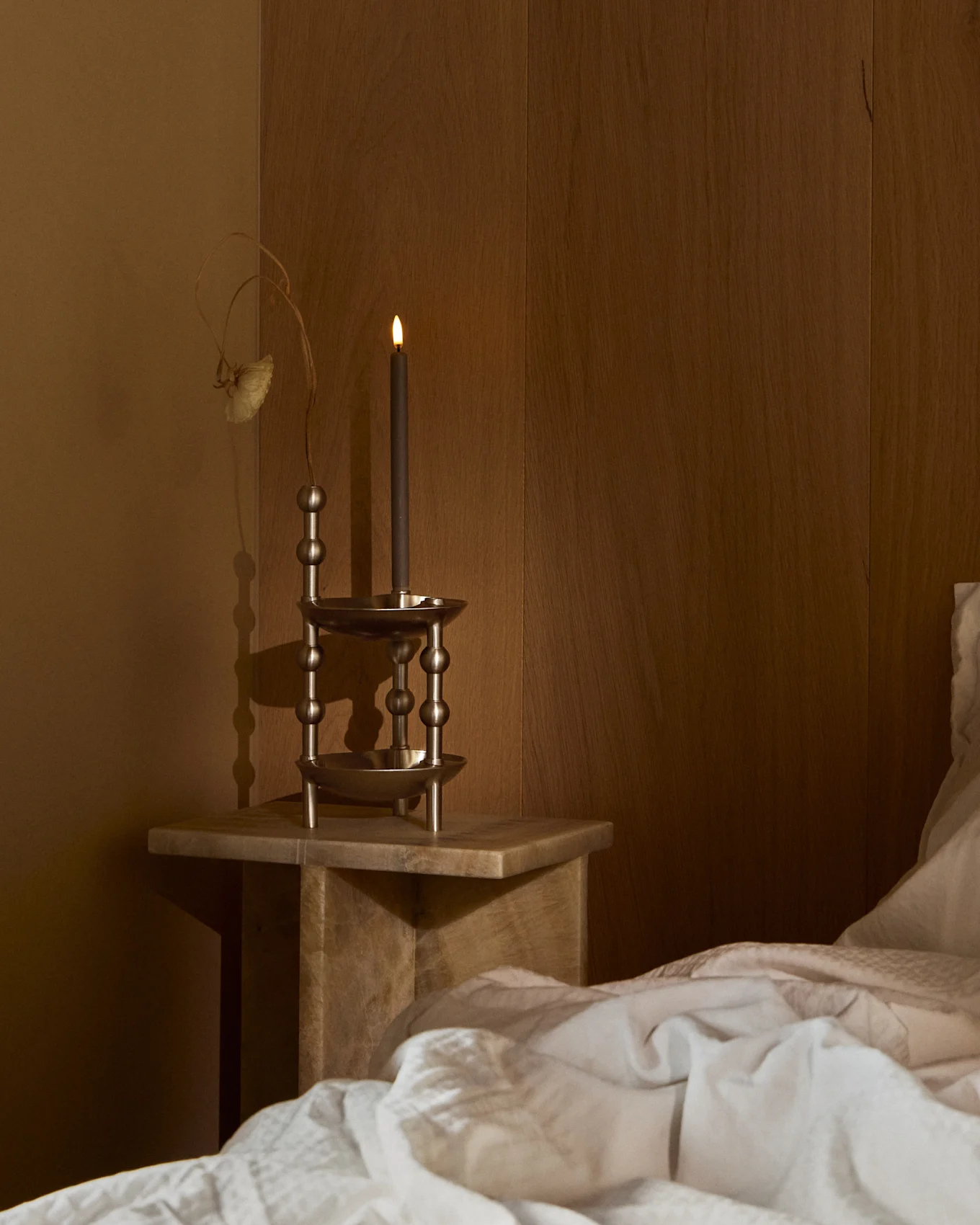 A lit dark candle in an ornate metal holder on a wooden bedside table next to a bed with white bedding in a dim room.