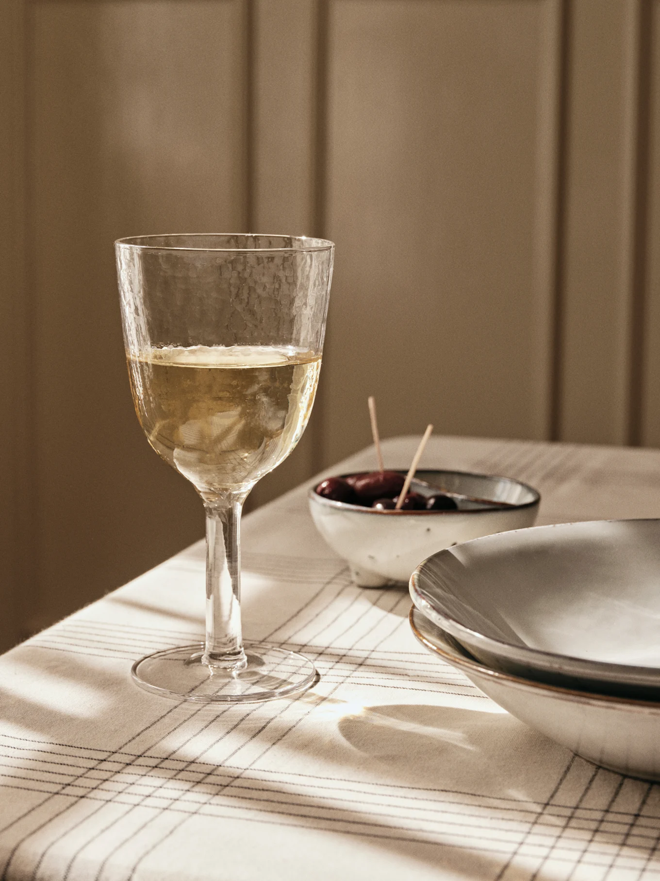 A textured glass of white wine on a striped tablecloth, with olives and plates in the background, bathed in window light.