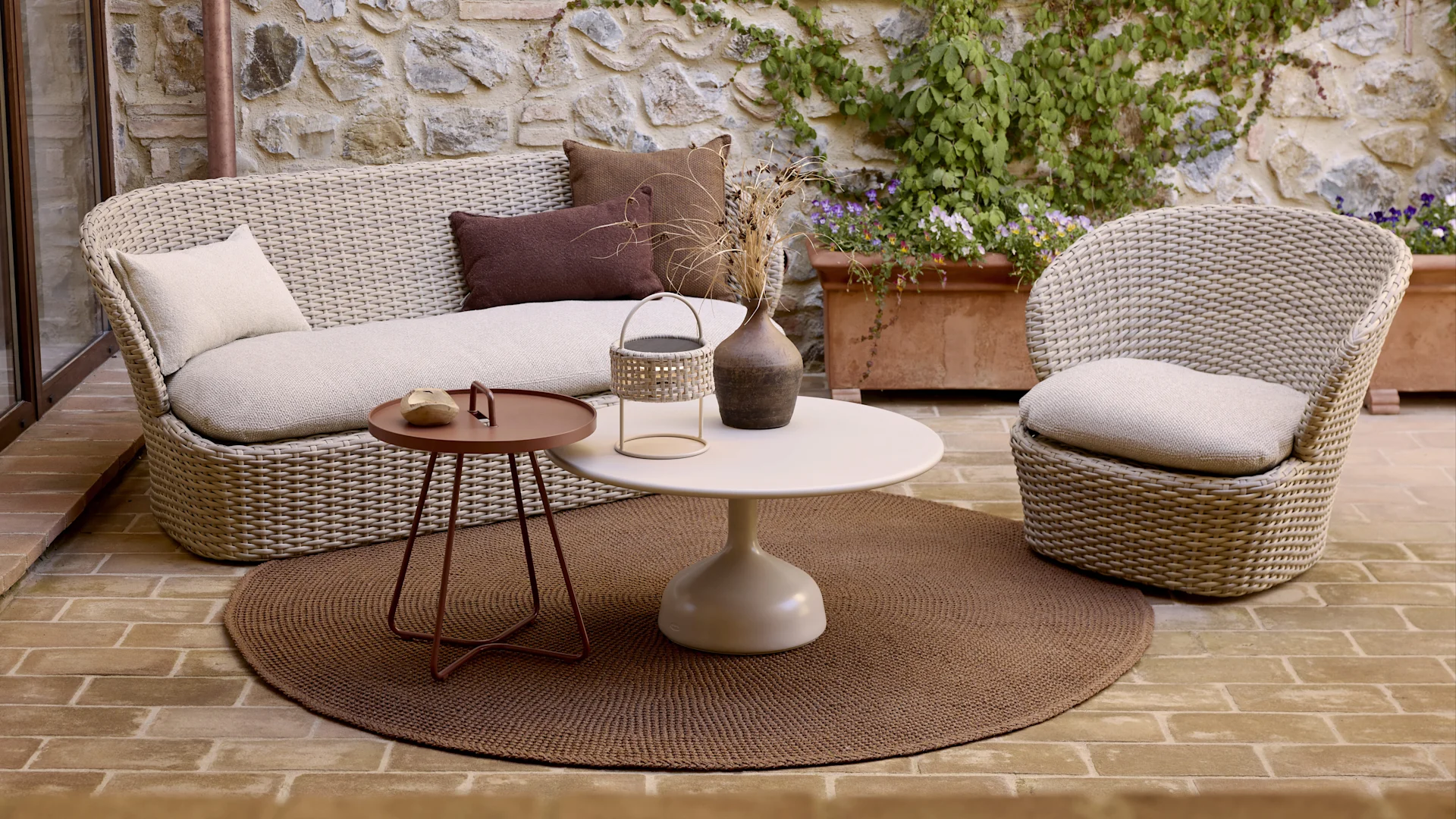 Outdoor lounge area with a woven sofa and armchair, round coffee table, side table, and a brown rug on a brick patio.