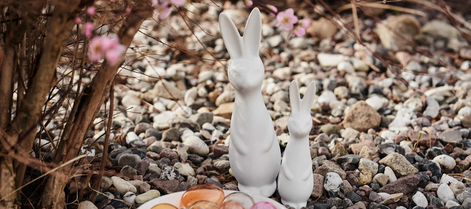 Two white ceramic Easter bunnies, a large and a small, stand among colorful pebbles with blurry pink flowers in the background.