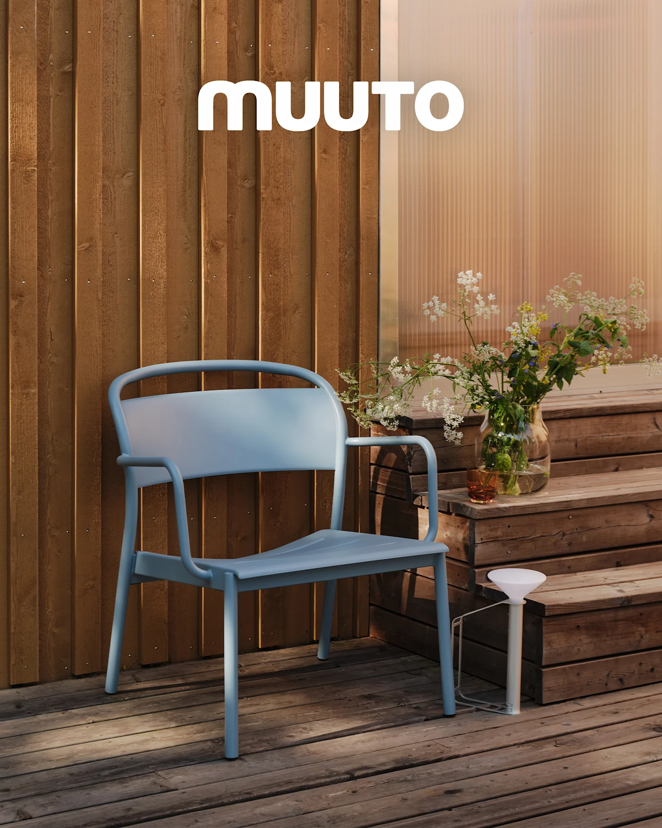 A light blue outdoor chair on a wooden deck next to steps with a vase of wildflowers and a white portable lamp. MUUTO logo.