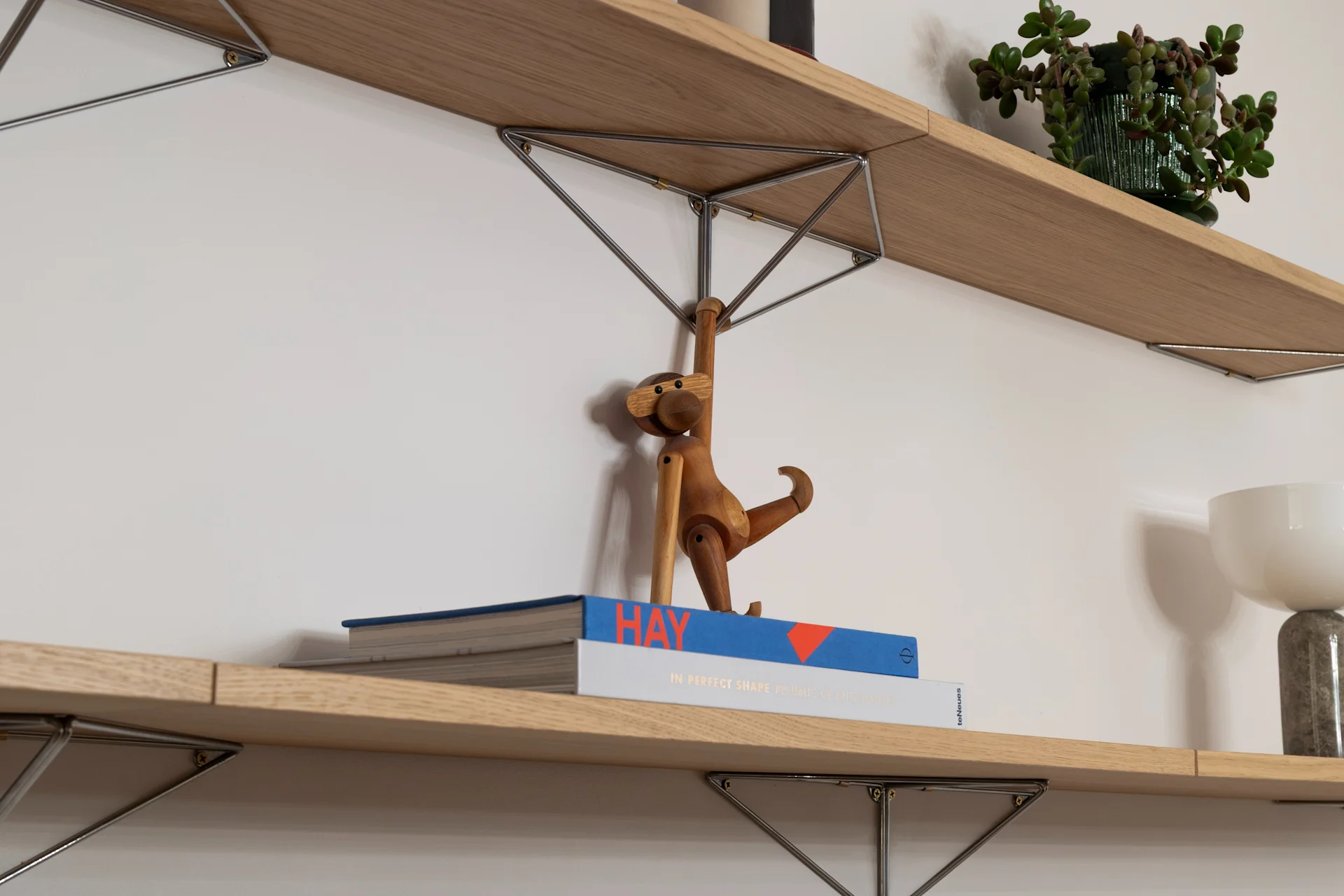 Two wood shelves on white wall. Top: striped candles, potted plant. Bottom: books, wooden monkey, white lamp.