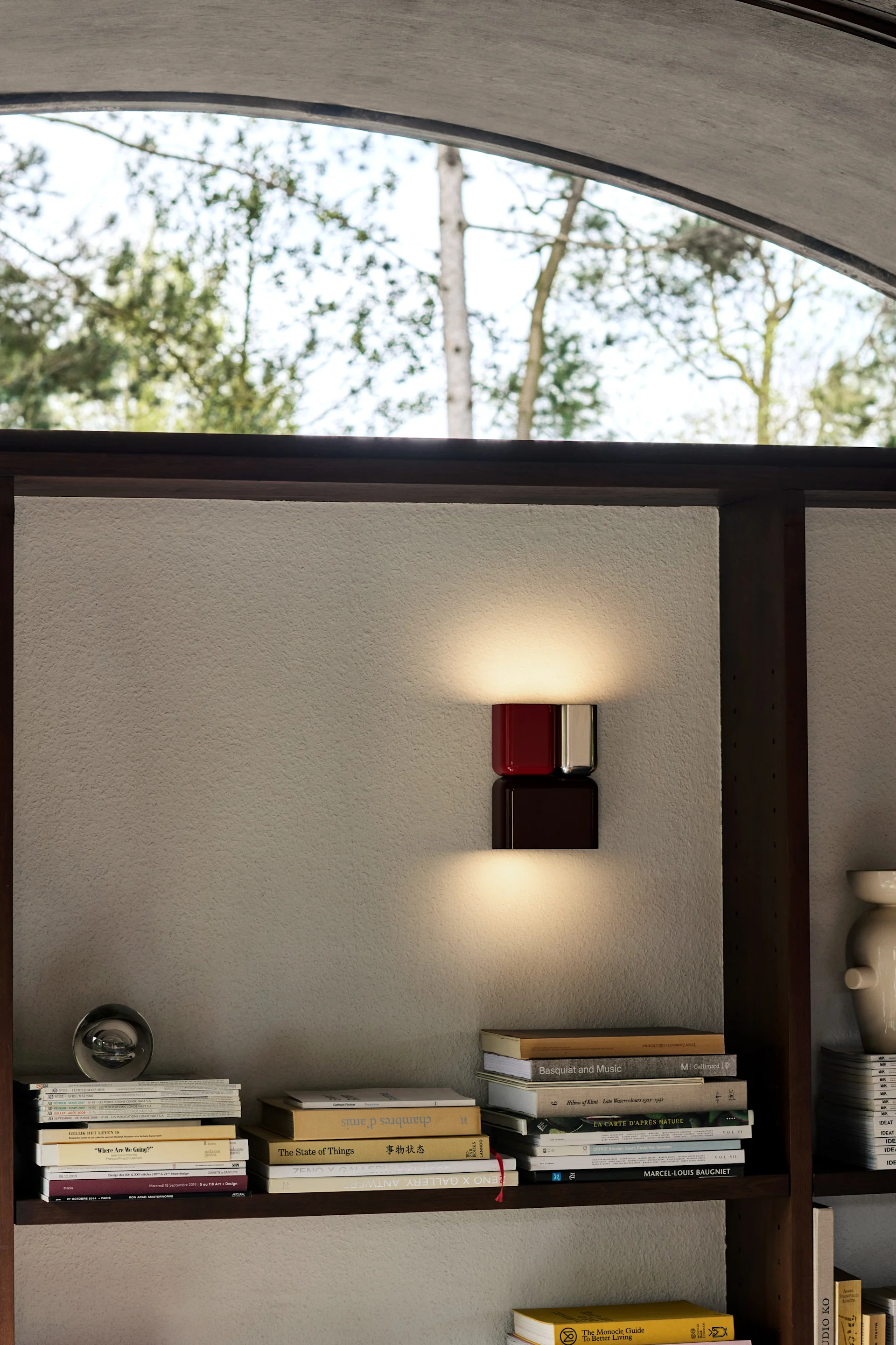 A modern wall sconce with a red upper and a dark brown lower cube-like element illuminates a textured wall between bookshelves filled with books.