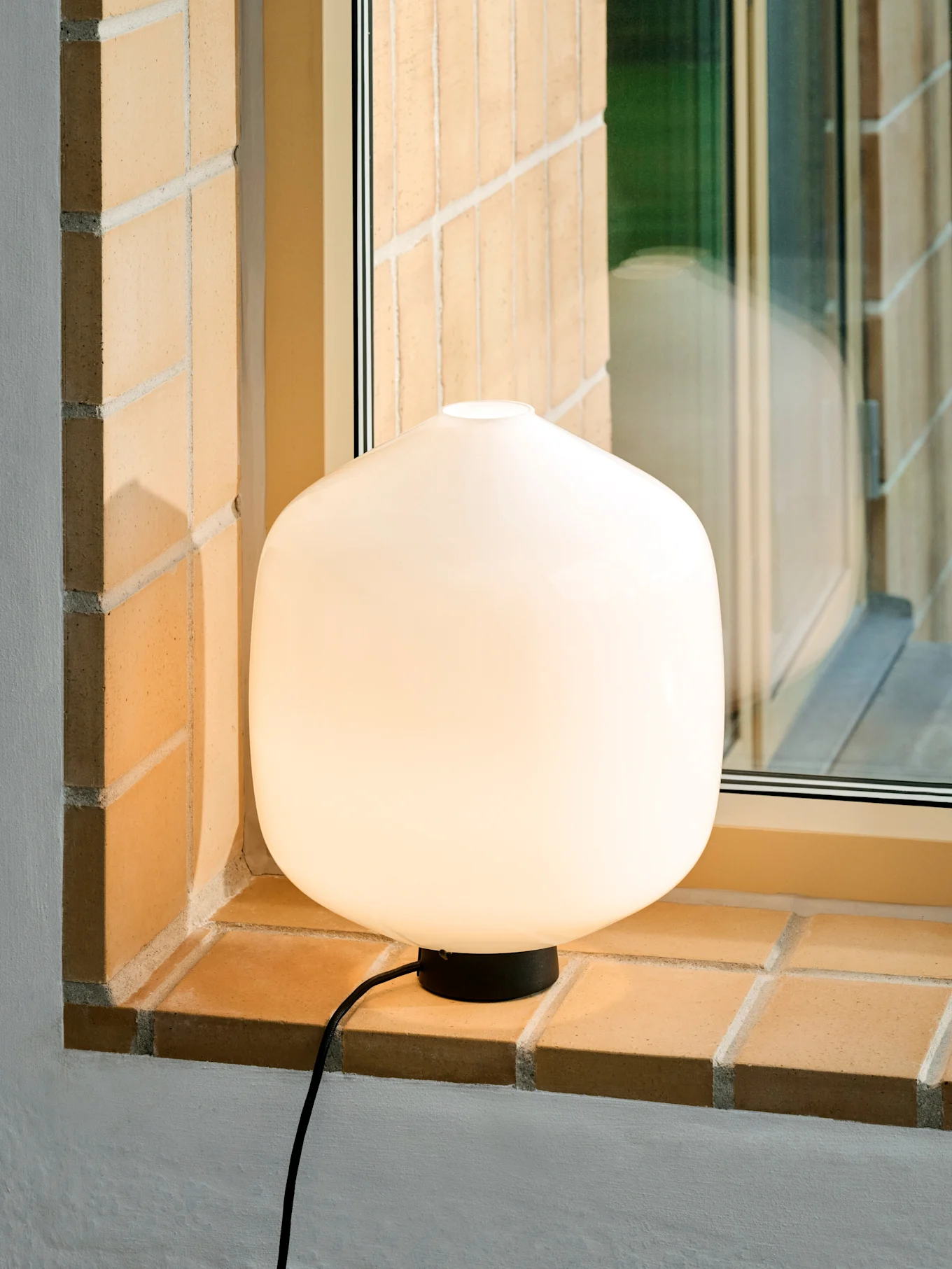 A glowing, rounded white table lamp with a black base and cord on a brick window sill.
