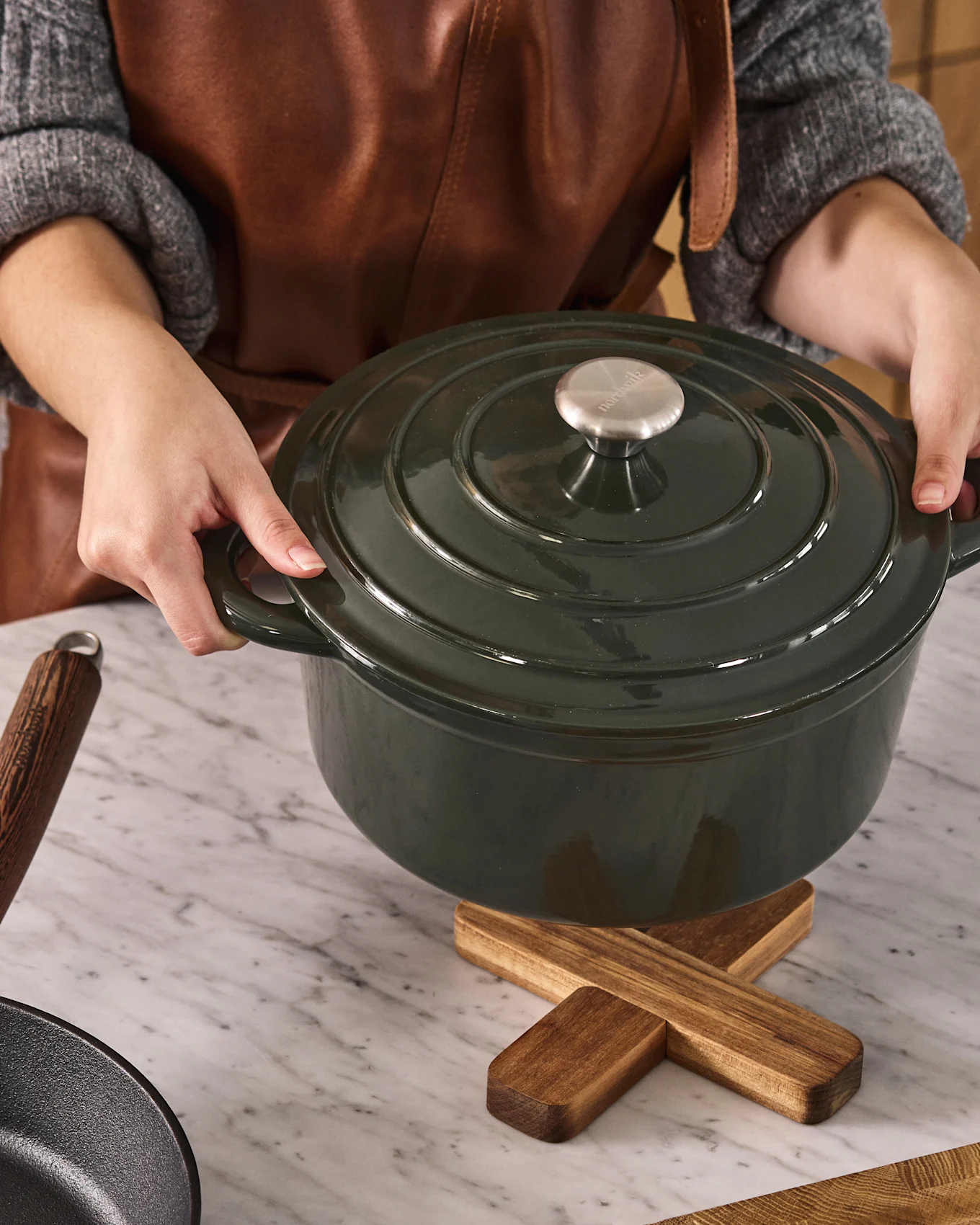 Hands in a brown apron and grey sweater hold a dark green enameled cast iron Dutch oven on a marble counter with a wooden trivet.
