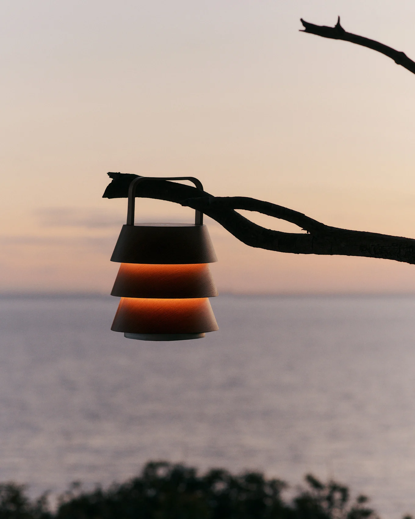 Lantern with tiered shades hanging from a branch against a sunset sky over water.