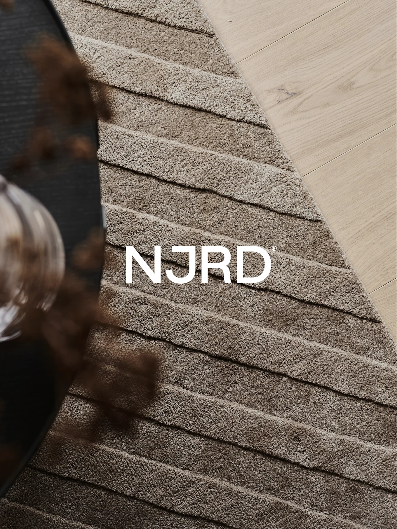 Close-up of a light brown shaggy rug with horizontal ridges, partially on a light wooden floor, featuring the white 'NJRD' logo.