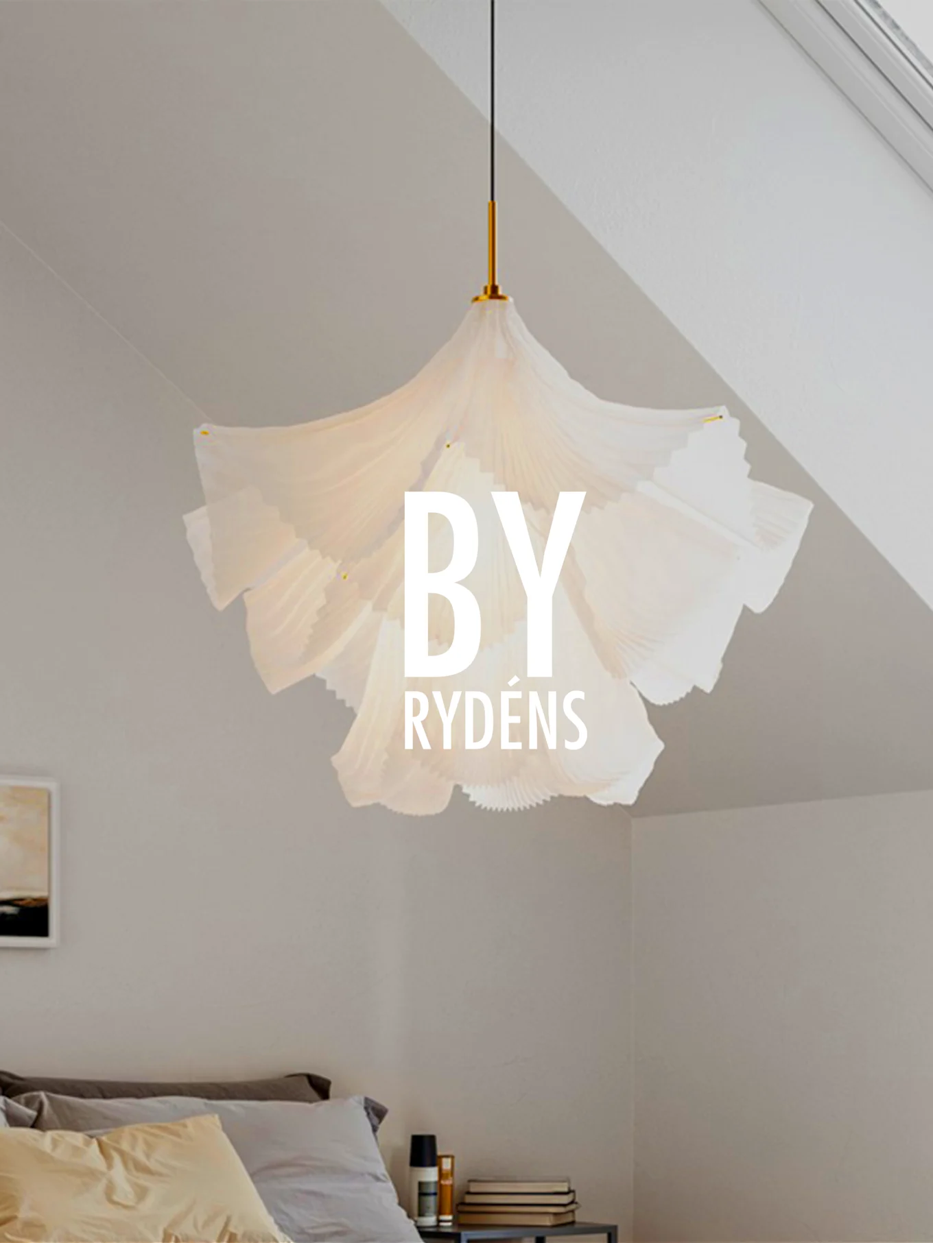 A white, pleated sculptural pendant lamp hangs above a bed with pillows and a bedside table. 'BY RYDÉNS' is overlaid on the lamp.