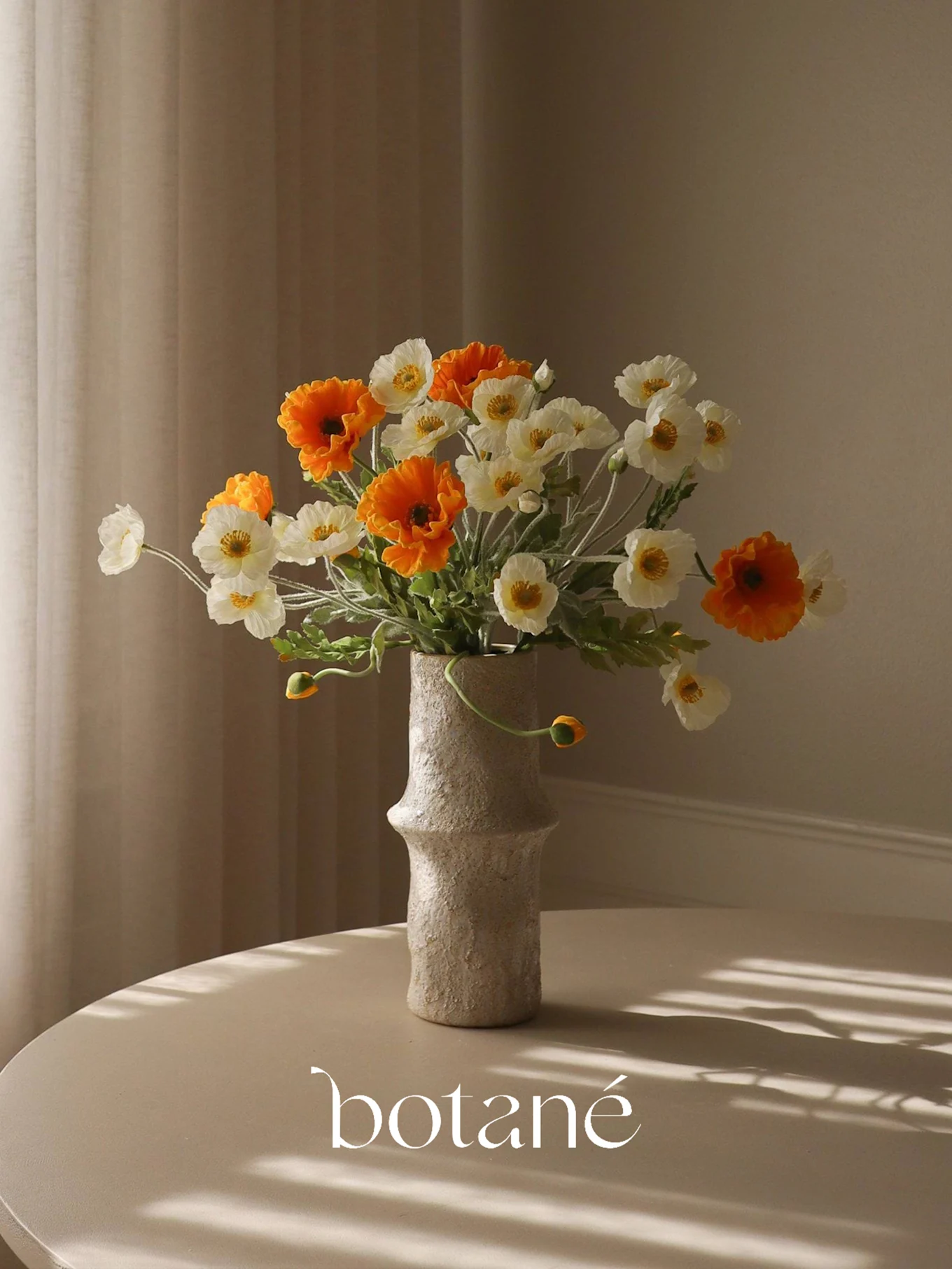 A textured vase holds a bouquet of orange and white faux poppies on a table with striped shadows.