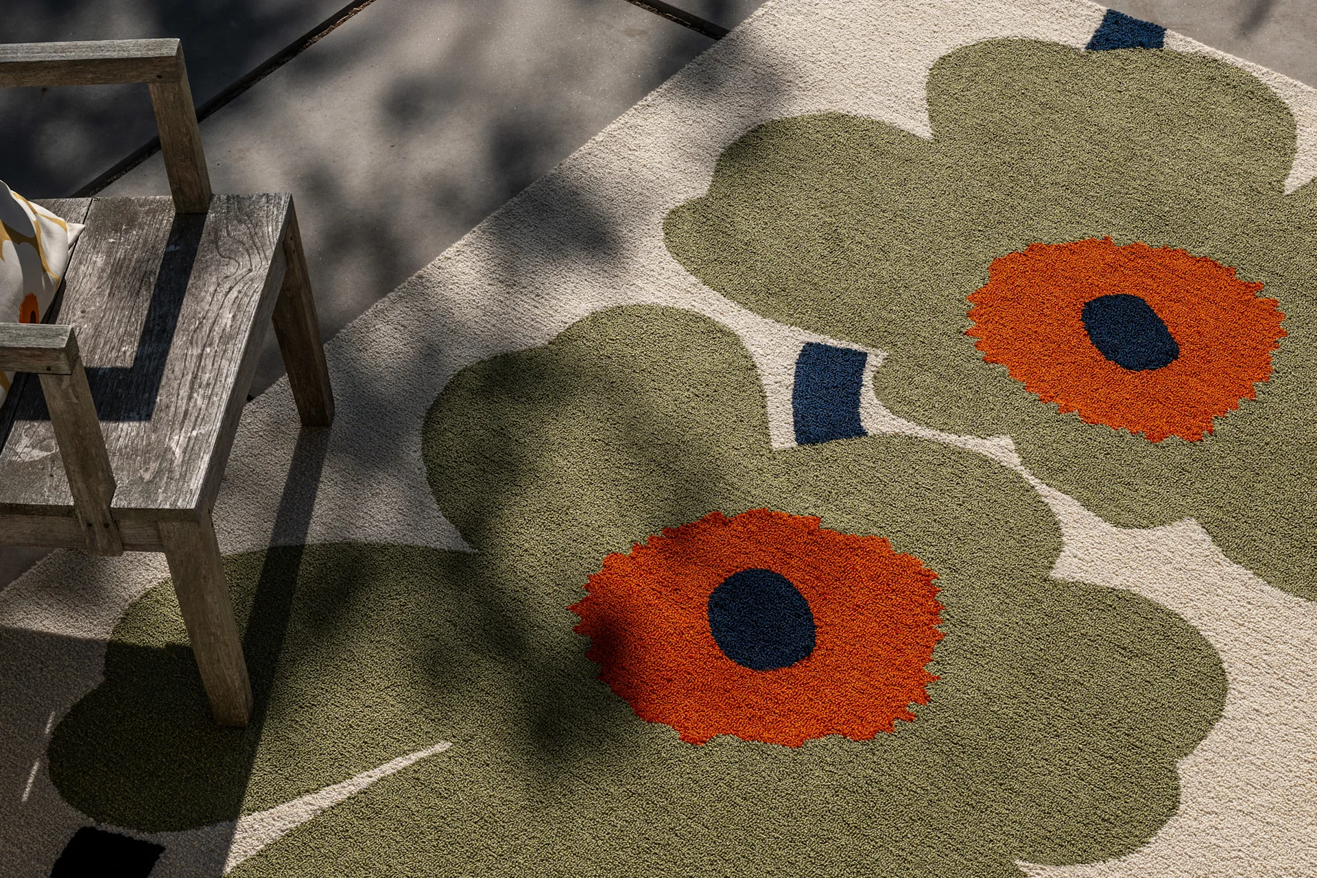 Outdoor rug with large olive green, orange, and dark blue floral pattern, next to a rustic wooden chair in dappled sunlight.