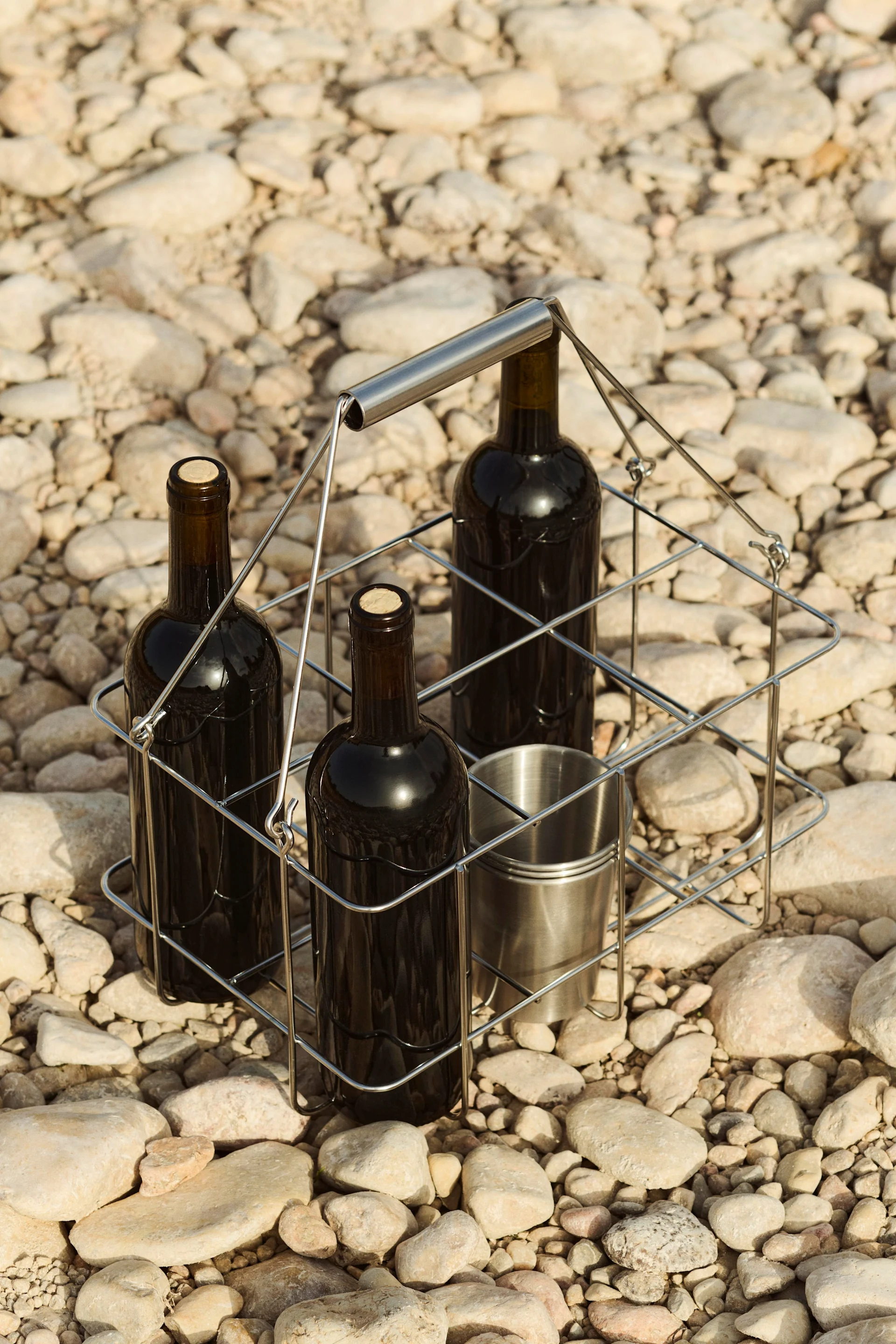 Three wine bottles and a metal cup in a portable carrier on a bed of pebbles.