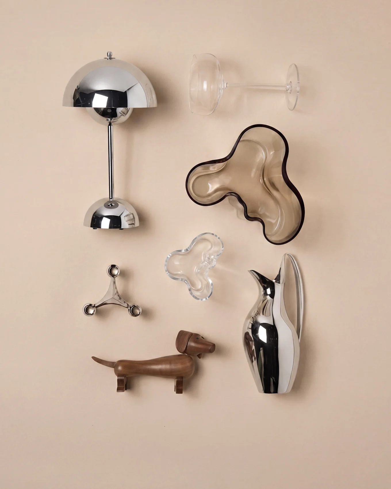 An arrangement of decorative objects: a chrome lamp, wine glass, two organic bowls, a candelabra, a wooden dachshund, and a pitcher.
