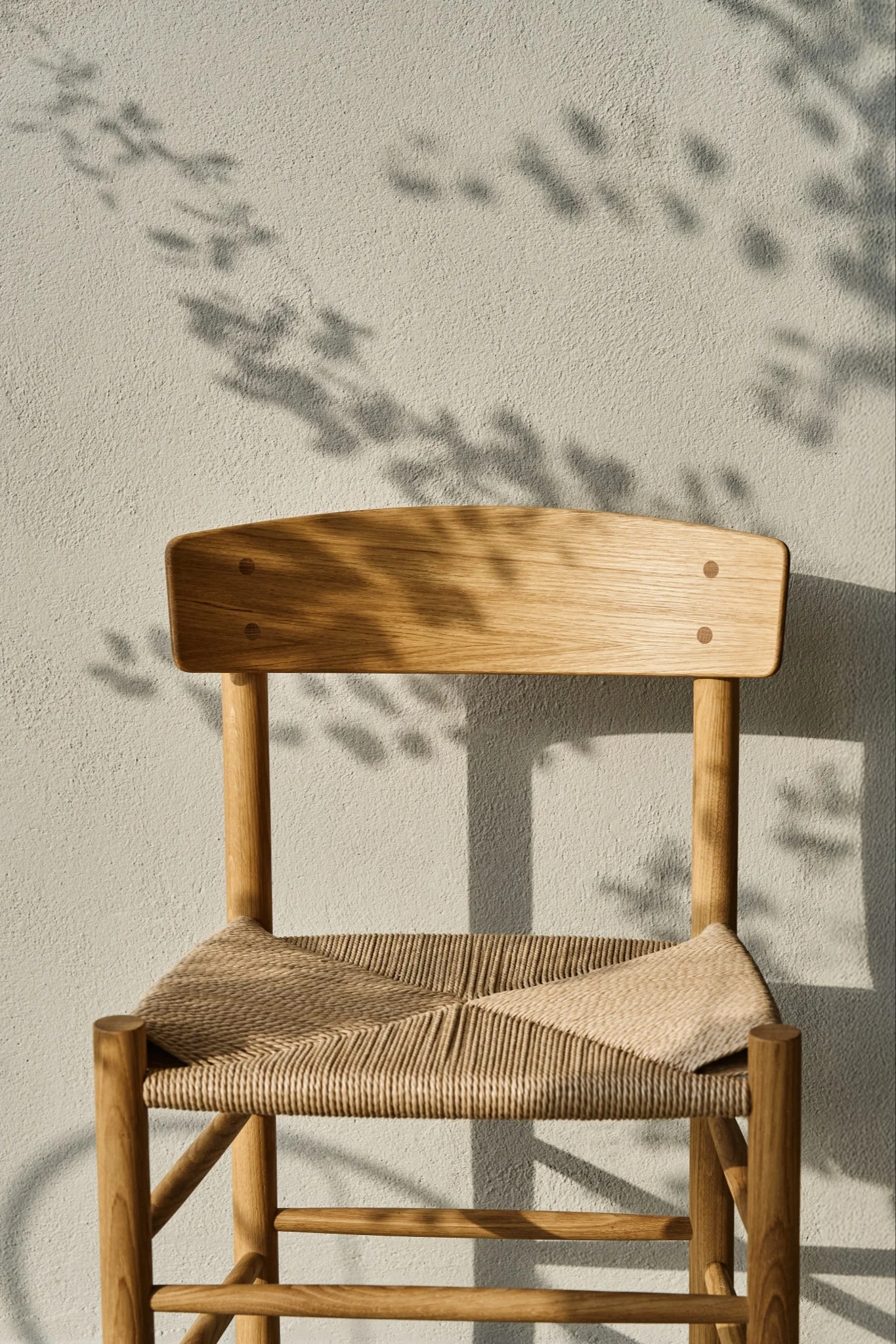 A light wood chair with a woven seat against a textured light wall, with leaf shadows.