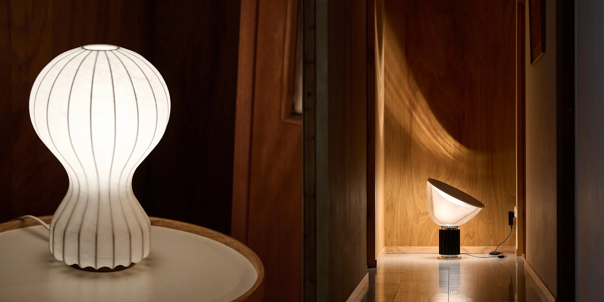 Two modern lamps; one a white ribbed table lamp, the other a conical floor lamp illuminating a wooden hallway.