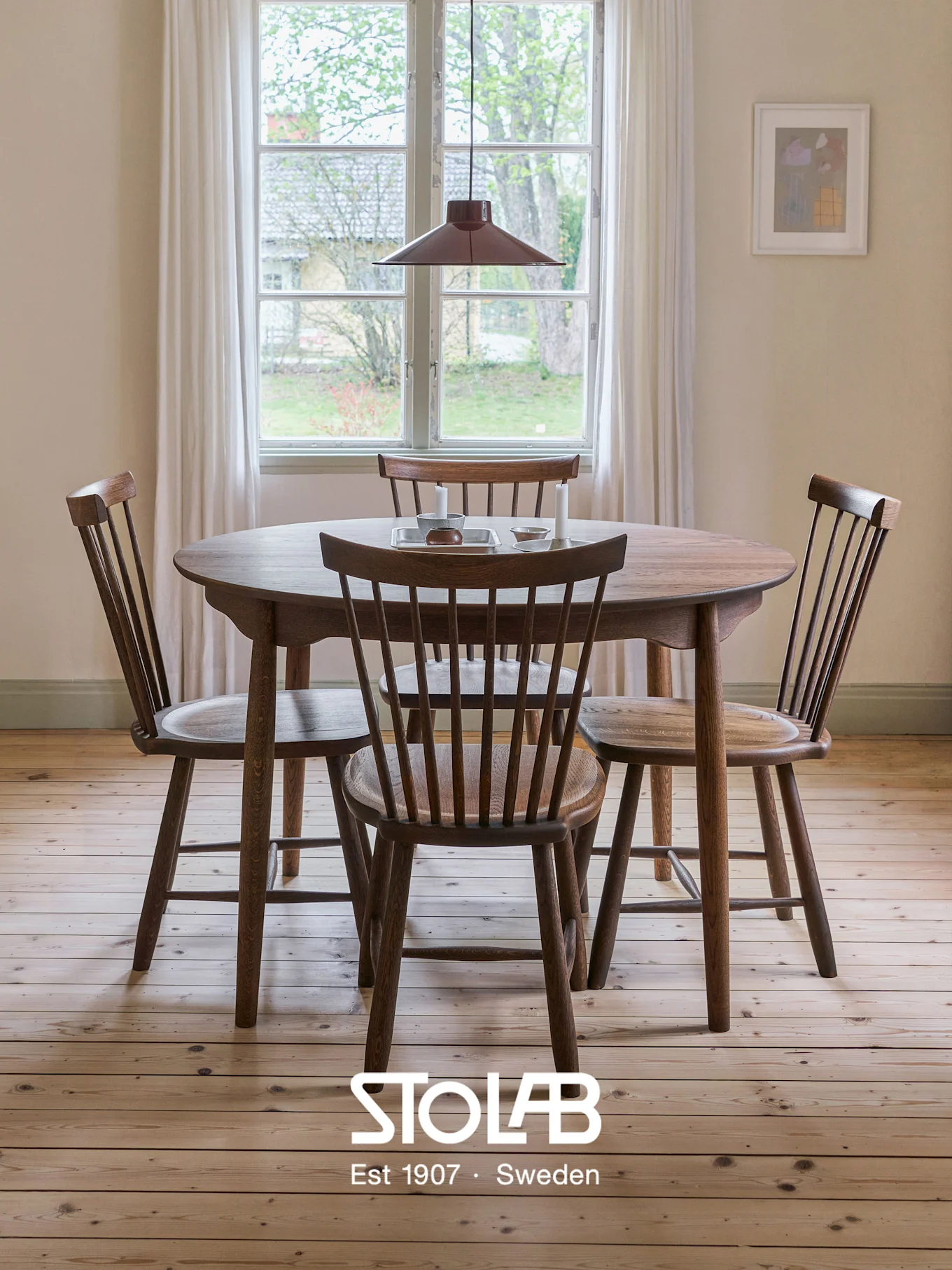 Round wooden dining table with four spindle-back chairs in a bright room, light wood floor, window, and red pendant lamp.