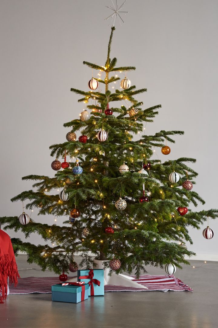 Decorate the Christmas tree with Christmas tree decorations for 2021 in 4 different styles according to Nest Trends - Nurture, Share, Boost and Cultivate. Here you see a colourful and playful Christmas tree on a striped rug from HAY in red and white and a white Christmas tree base from Born in Sweden.