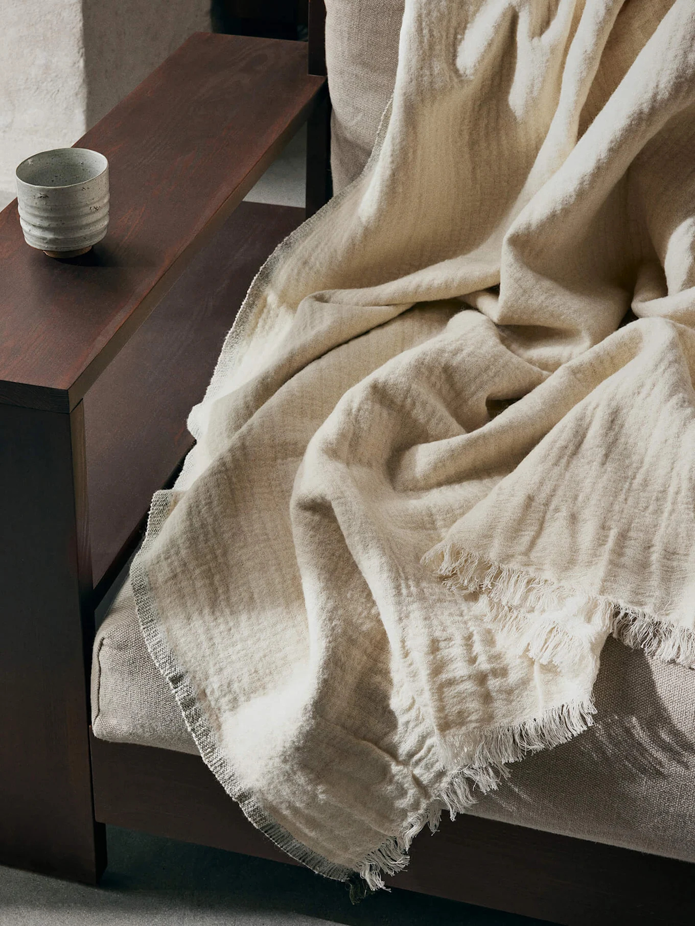 A light beige textured blanket with frayed edges draped over a sofa, with a ceramic cup on a dark wooden armrest.