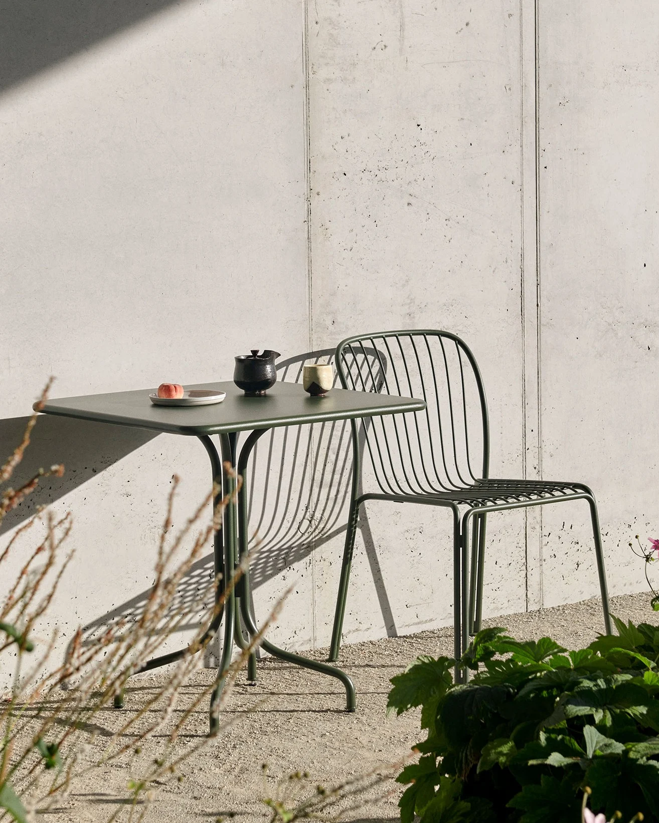 A dark green outdoor table with a peach, teapot, and cup, and a matching slatted chair, against a concrete wall with strong shadows.