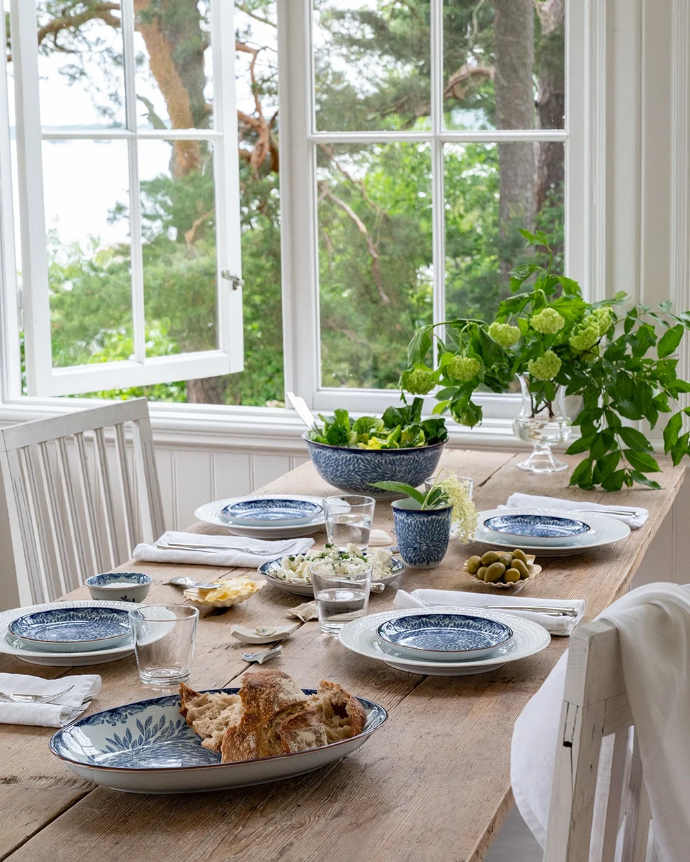 A rustic wooden dining table set with blue and white dishes, salad, bread, and olives, by an open window overlooking green trees and water.