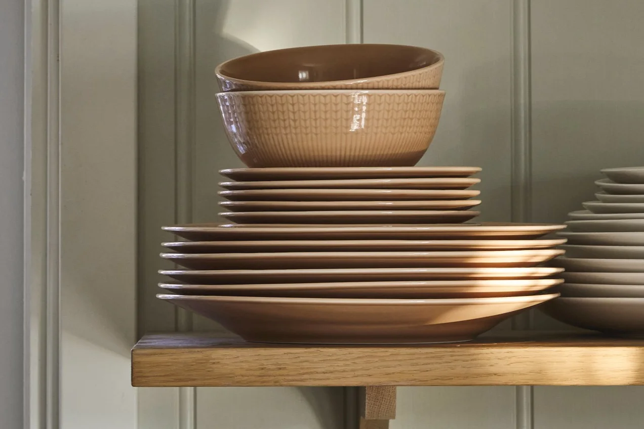 Stacks of light brown ceramic plates and patterned bowls on a wooden shelf.