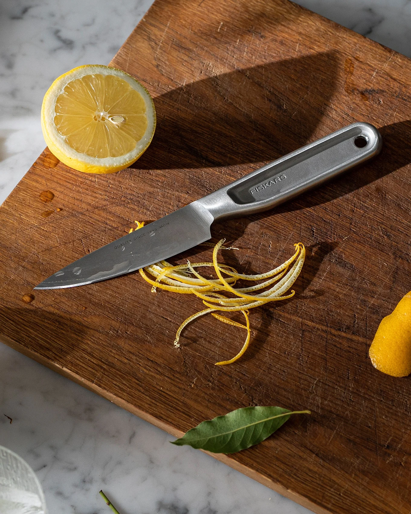A stainless steel knife on a wooden cutting board with lemon halves, lemon peels, and bay leaves on a marble counter with a glass of ice.