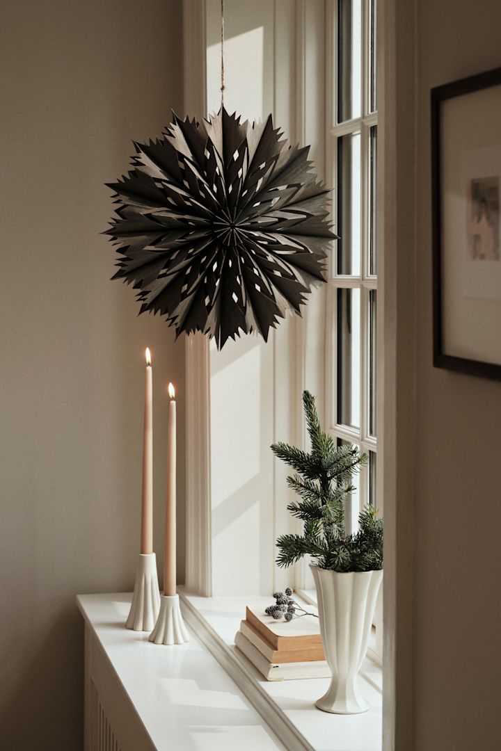 The paper star lantern for Christmas - Venja - from Broste Copenhagen hangs in the window with a pair of candlesticks and a vase. 