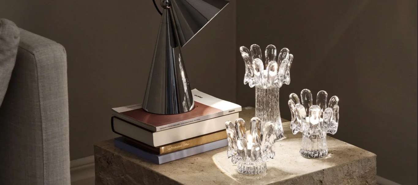 Three glass candle holders in different sizes sit on a stone table alongside a portable lamp and books, together reflecting the Scandinavian interior trends design trends for 2026.