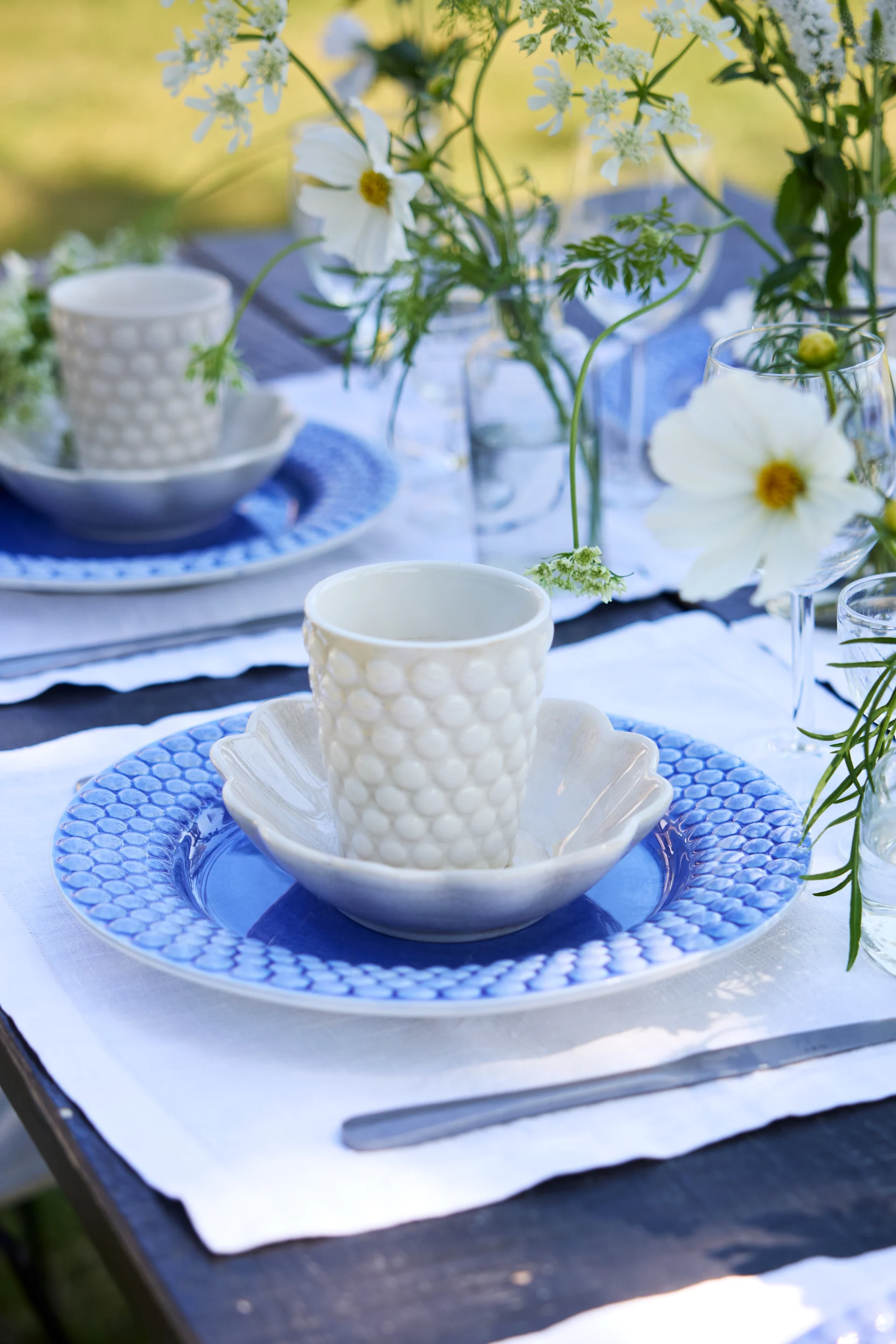A close-up of an outdoor table set in blue and white. A blue Bubbles plate together with a white Bubbles cup on a table decorated with flowers.