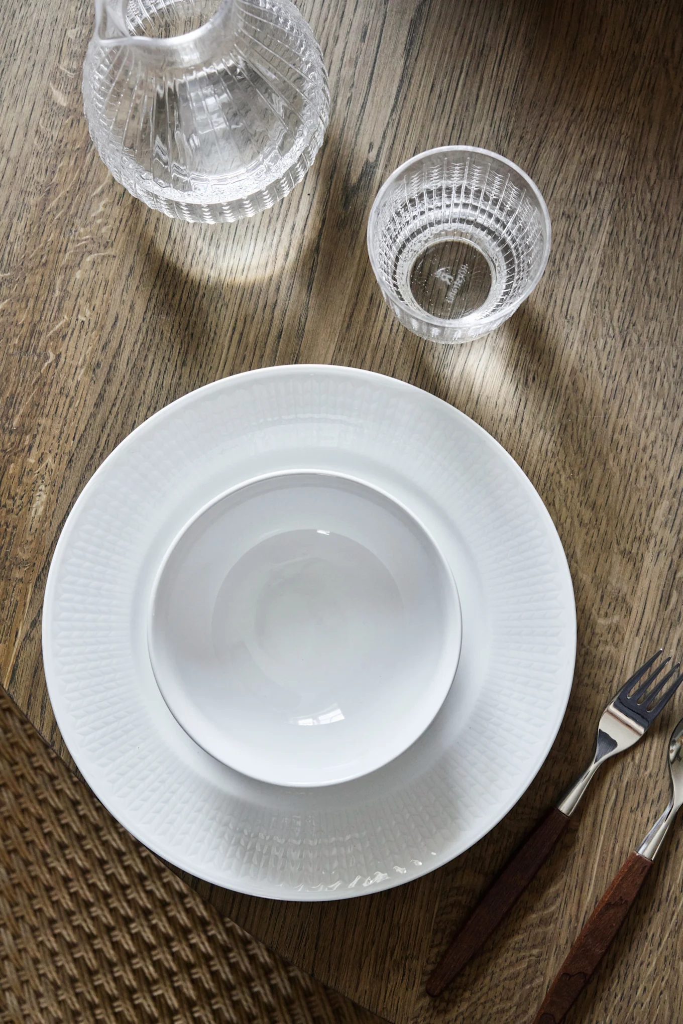 Overhead shot of a minimalist table setting with white plates, a glass carafe, a glass, and cutlery on a wooden table.
