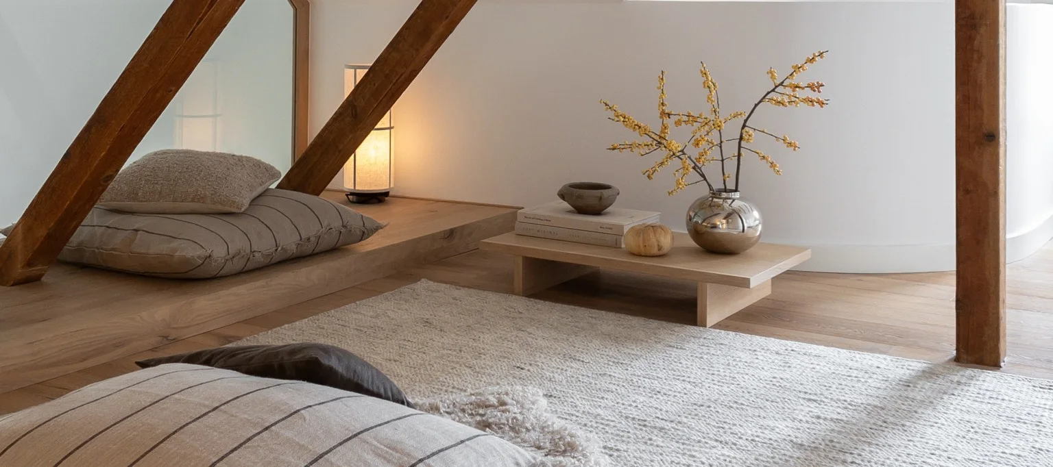 A serene, minimalist room featuring light-colored floor cushions, a woven rug, and a low wooden table holding a silver vase with yellow berries, books, and a lamp.