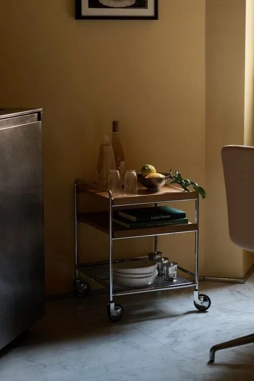 A three-tiered utility cart with metal frame and wooden shelves, holding bottles, glasses, lemons, books, and plates.