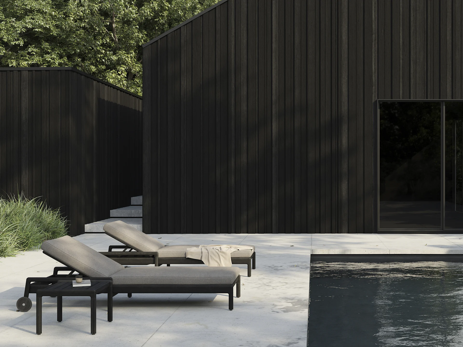 Two dark sun loungers with light cushions on a patio next to a swimming pool, with modern dark wood buildings in the background.