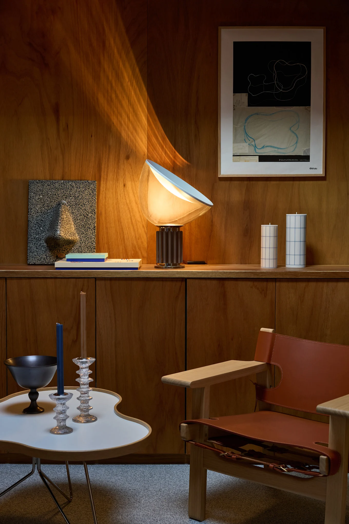 A living space with wooden walls, a glowing table lamp, abstract wall art, decorative candles, a modern side table, and a leather armchair.
