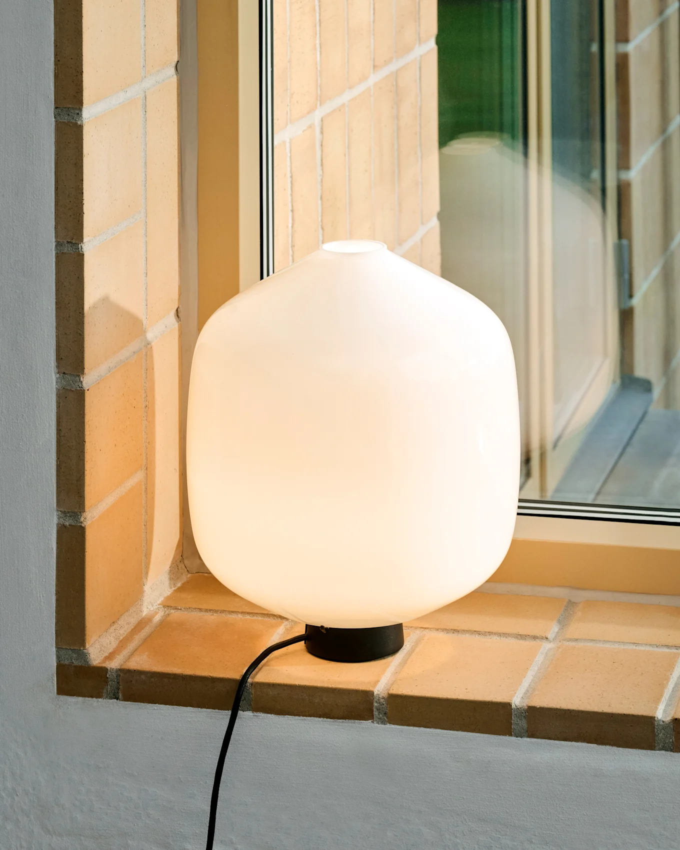 A glowing white rounded table lamp with a black base and cord sits on a light brick window sill.