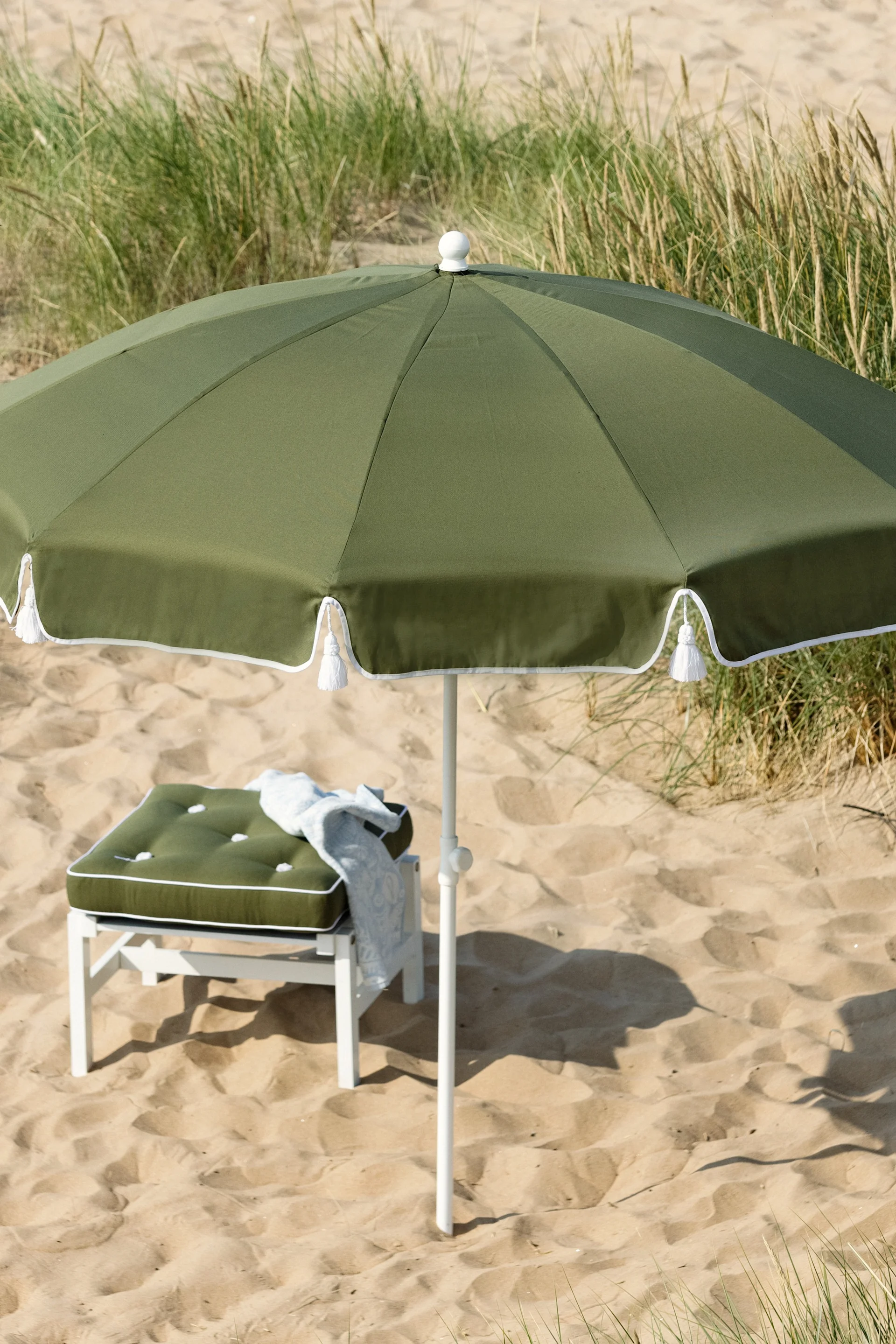 Olive green beach umbrella and matching cushion on a white stool on a sandy beach with green grasses.