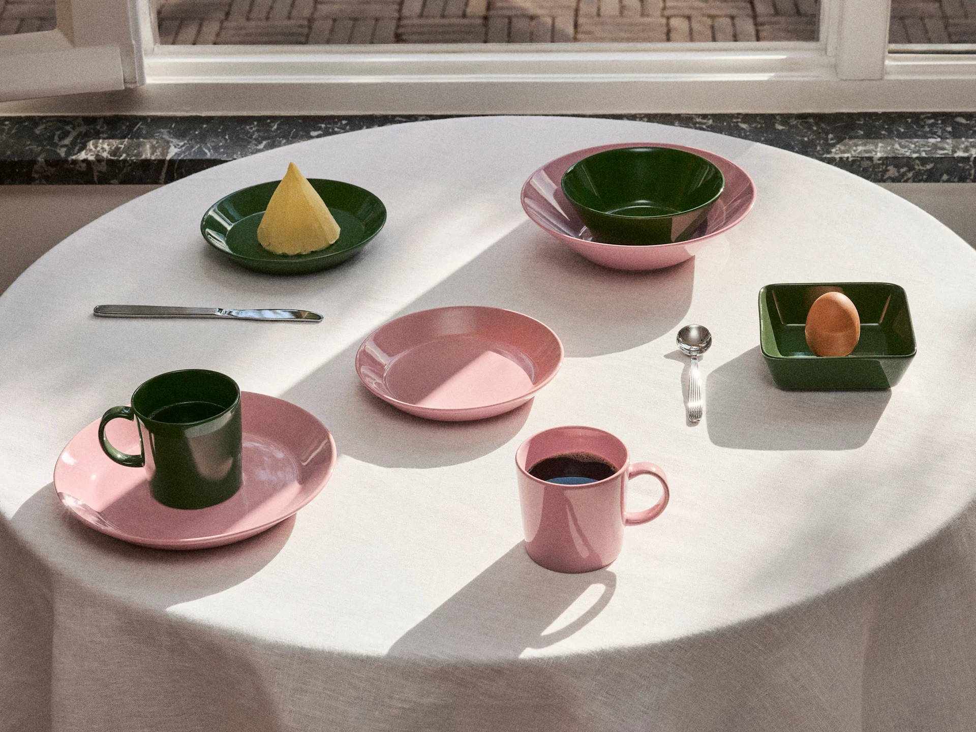Table set with pink and dark green tableware, including plates, bowls, and mugs, with butter, an egg, and coffee under natural light.