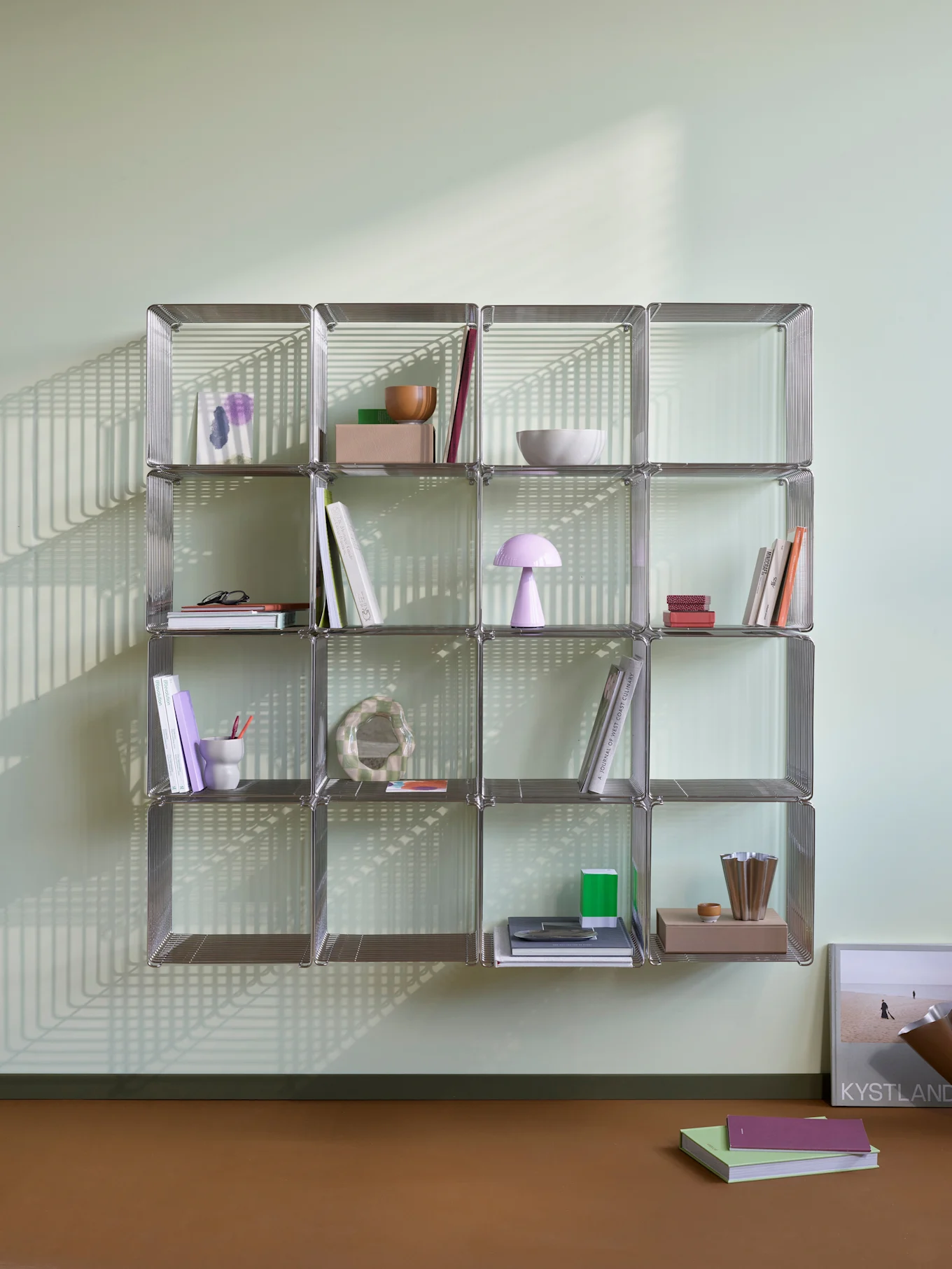 A chrome-like modular shelving unit with books and decorative items, illuminated by strong linear shadows on a light green wall and brown floor.