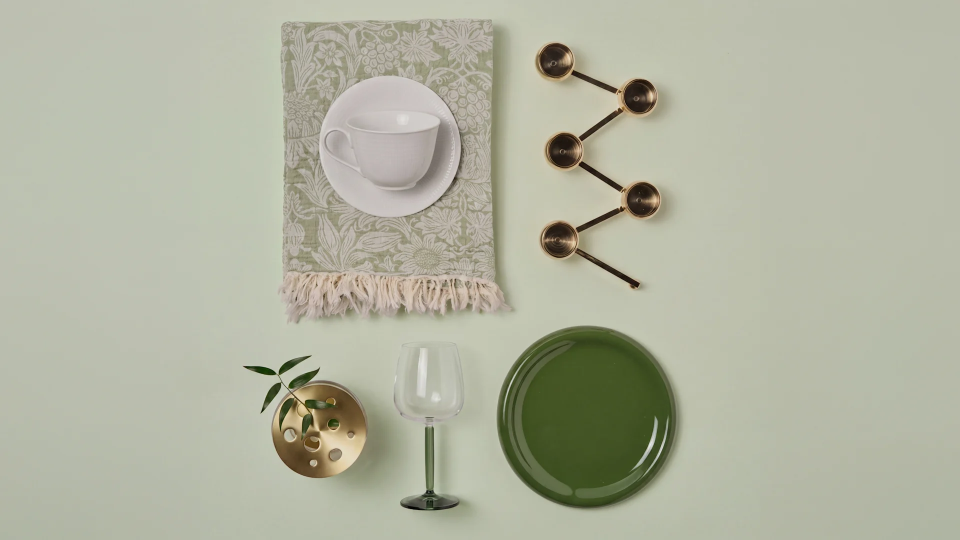 Flat lay: White teacup on green patterned napkin, brass measuring spoons, brass plant vase, green wine glass, dark green plate.