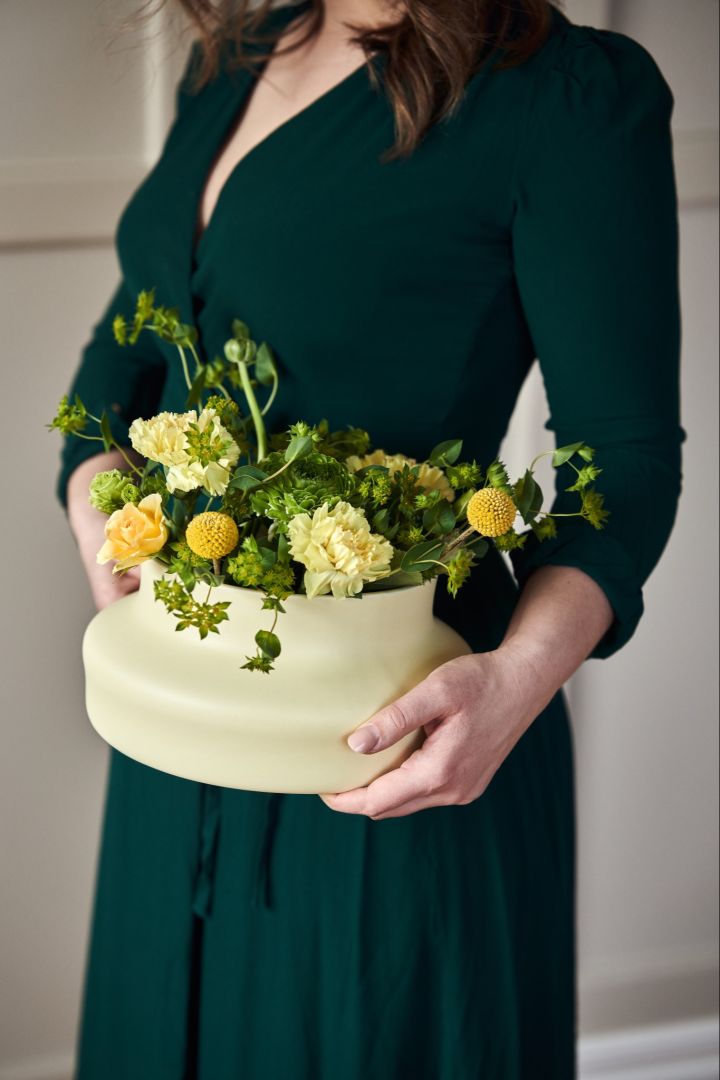  A woman in a green dress stands holding the mellow yellow low ceramic vase from the Dorotea collection for Gense. 