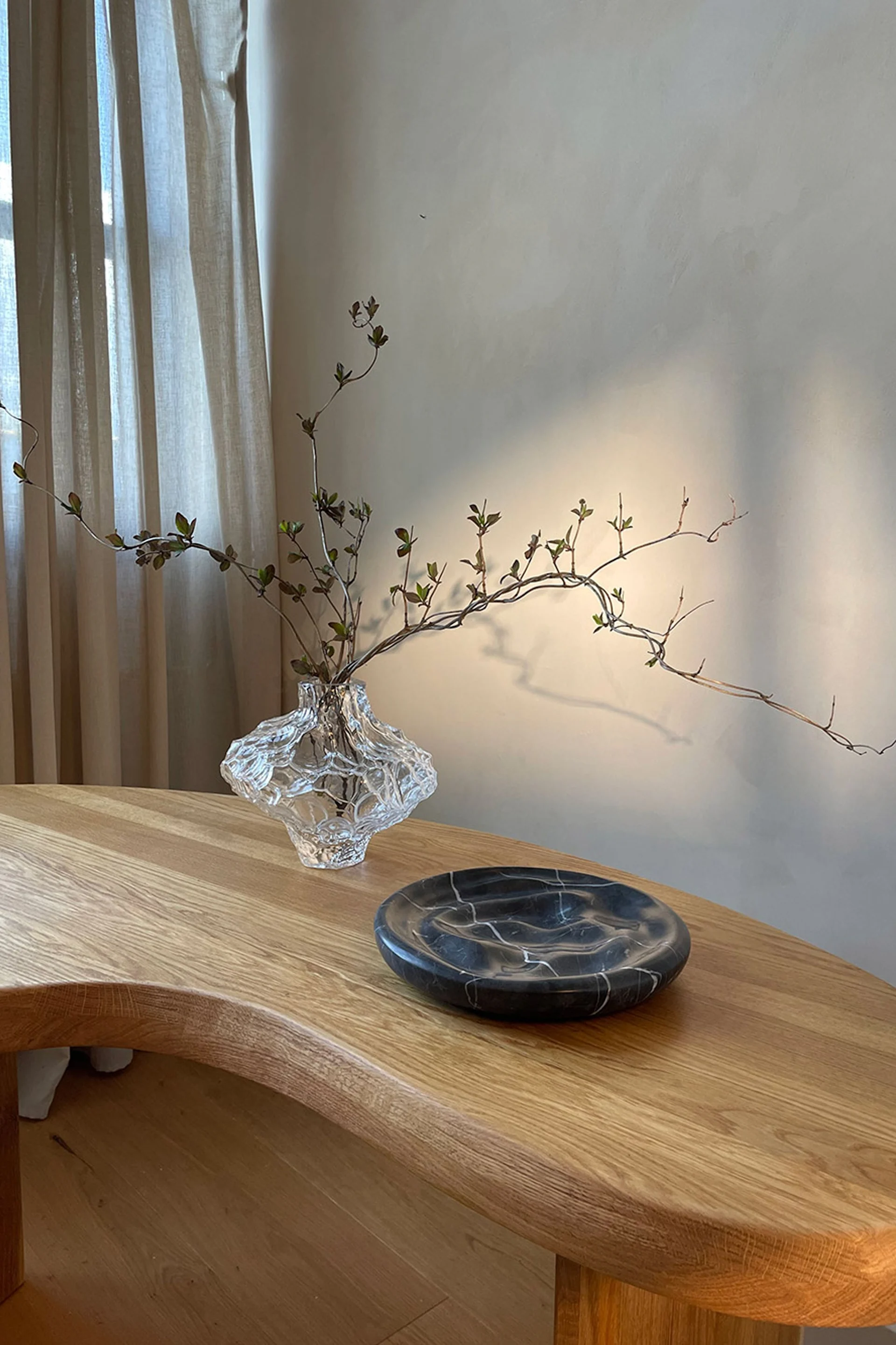 Clear textured glass vase with leafy branches and a dark marble plate on a light wooden table.