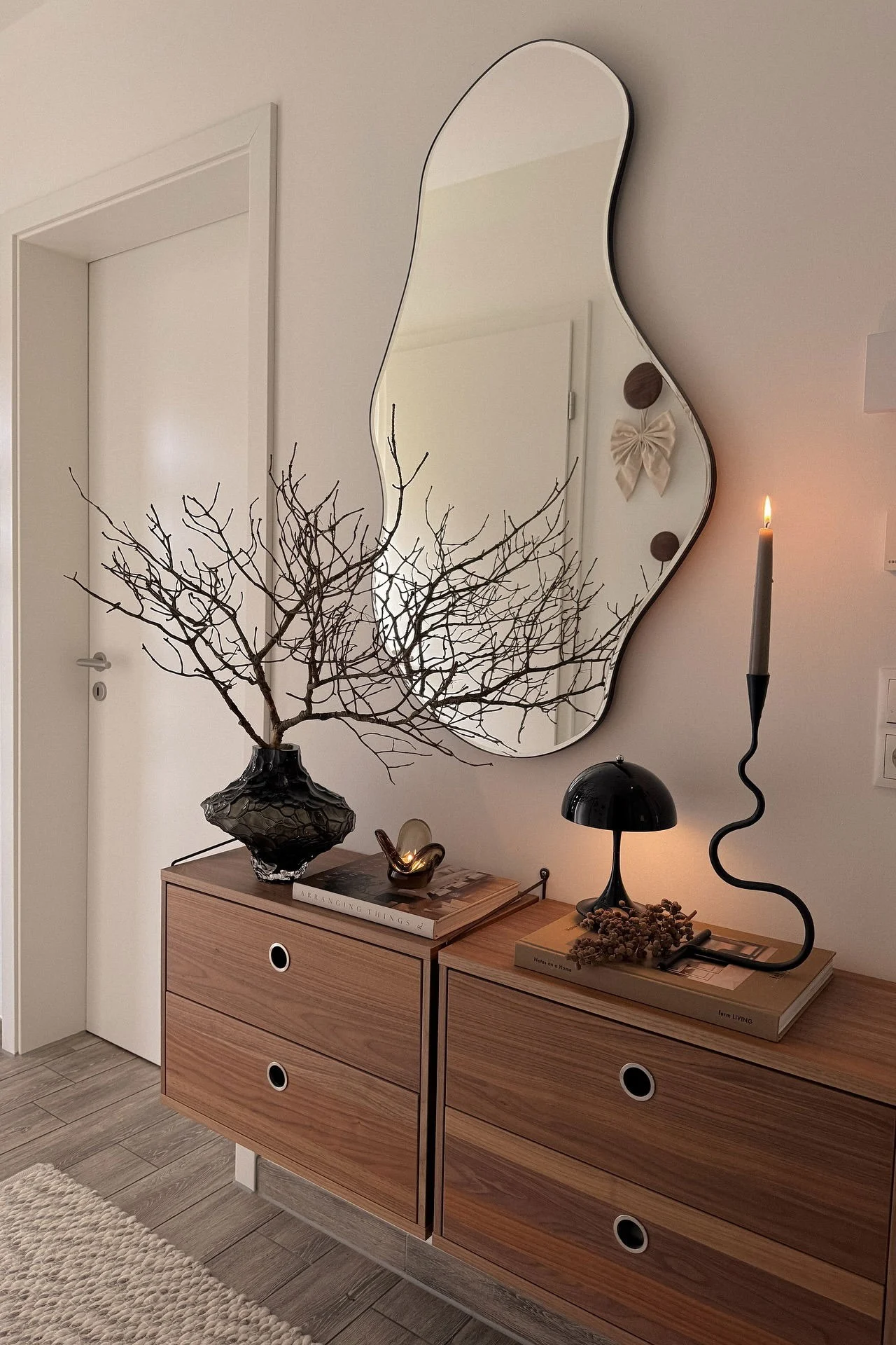 A console table with a dark vase of bare branches, a wavy candle holder with a lit candle, a black lamp, and an irregular mirror.