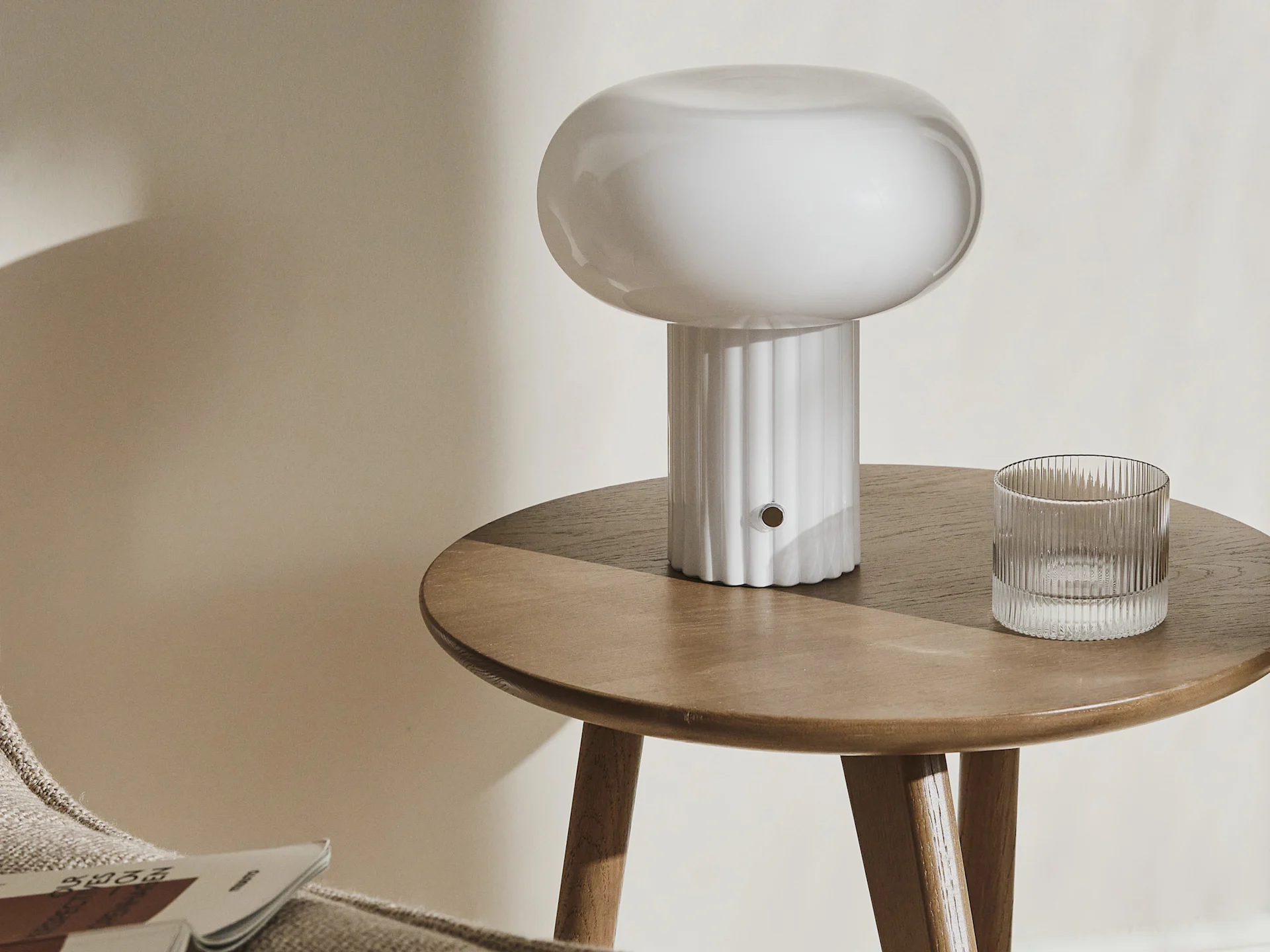A white mushroom lamp and a ribbed glass tumbler on a round wooden side table.