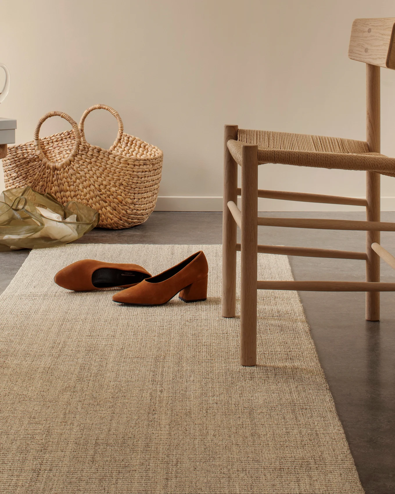 Brown suede block-heel shoes on a light woven rug, with a straw bag and a wooden chair in the background.